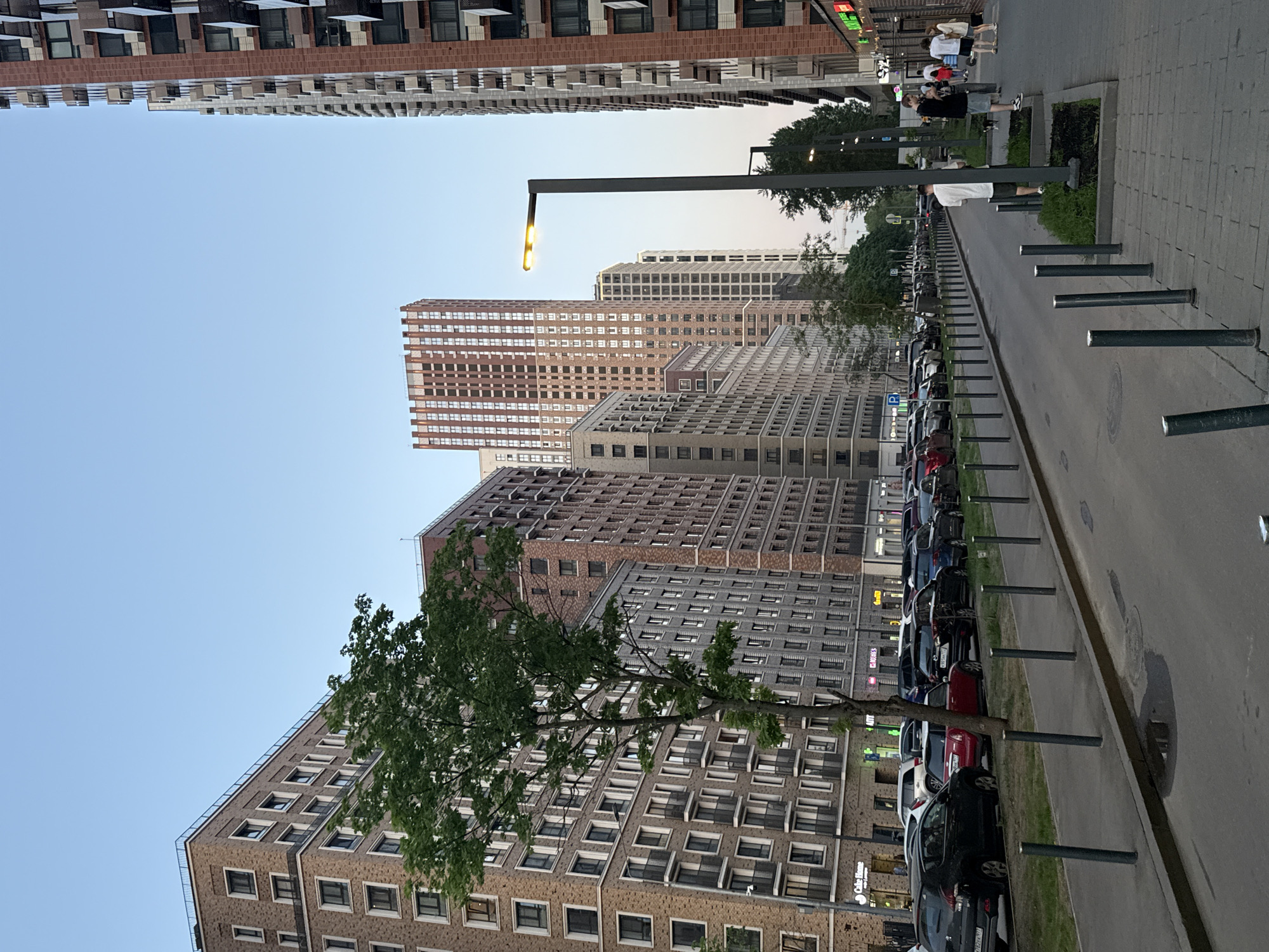 The image depicts a street scene in Moscow, Russia. Tall, modern apartment buildings line both sides of the street. Numerous cars are parked along the right side of the road, between metal bollards that separate the parking area from the sidewalk. A young tree stands near the parked cars.
On the right sidewalk, several people are walking. A lamppost stands tall, casting light. The sky is a pale blue, indicating either early morning or late evening. The overall environment is urban and contemporary.