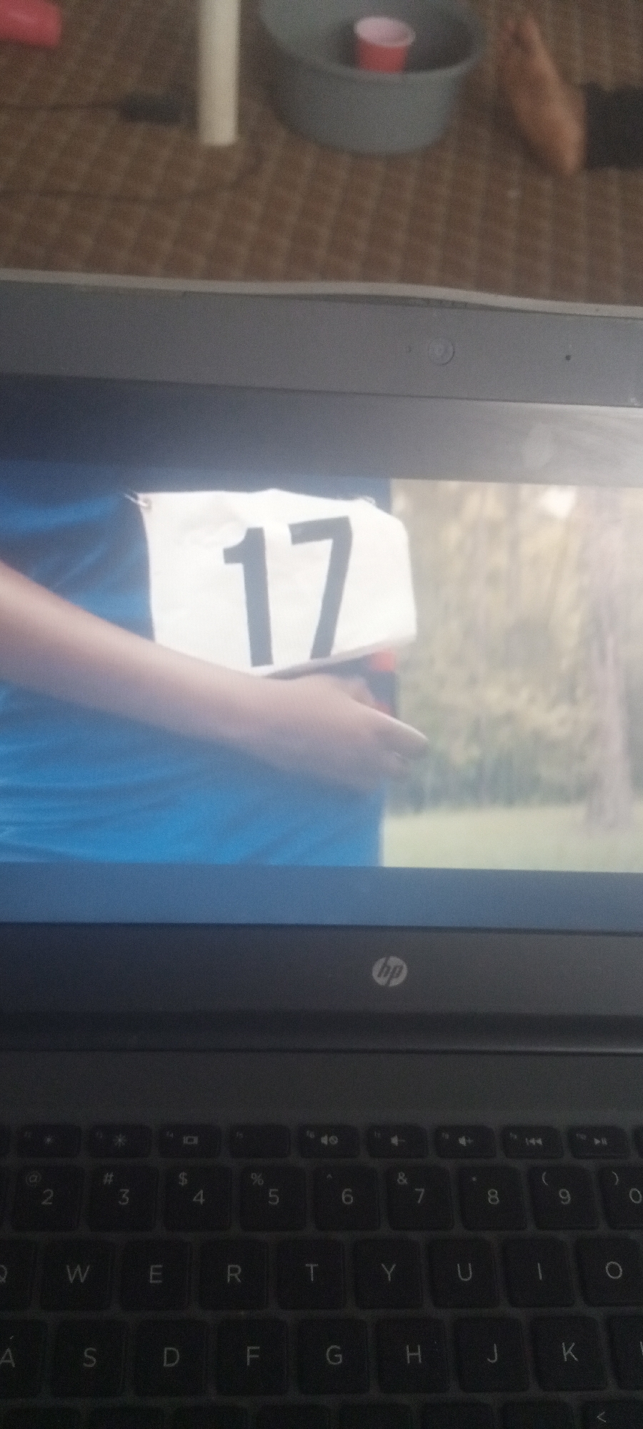 The image shows a laptop displaying a person wearing a blue shirt and a white tag with the number "17" printed in black. The person's arm and hand are visible as they hold the tag. The background of the laptop screen appears to be a blurred outdoor scene with trees. The laptop is open, and the keyboard and HP logo are visible. The top part of the image shows a glimpse of a room setting with a gray container, a red cup inside the container, and a brown patterned floor. The location is unknown.