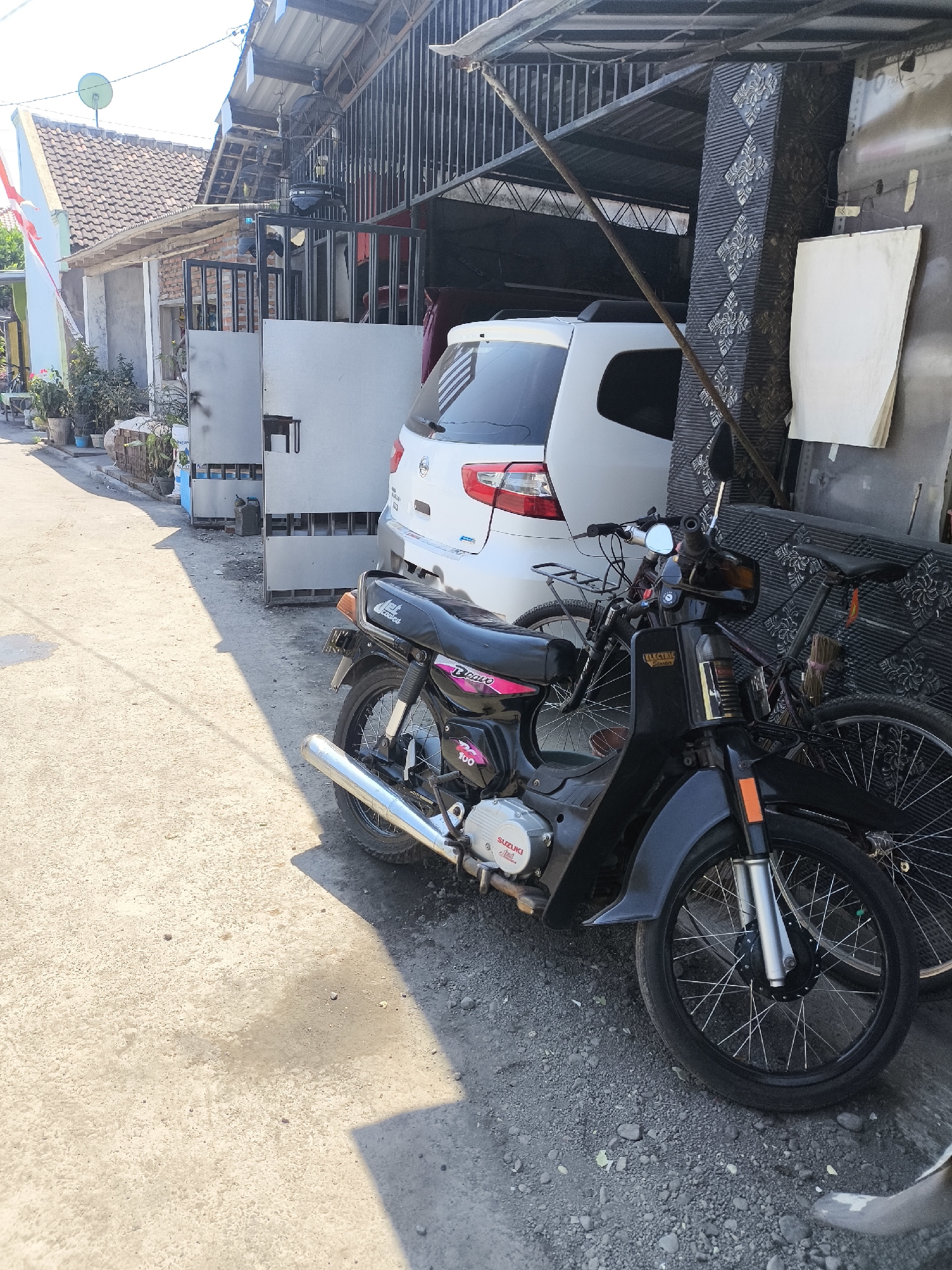 In the city of Lumajang, Indonesia, a black motorcycle is parked on a gravel and concrete surface in front of a house with a white car partially visible behind it. The motorcycle is a classic model with a prominent exhaust pipe, silver engine casing, and a black seat with pink accents. A bicycle is also parked behind the motorcycle. The scene is illuminated by sunlight, casting shadows on the ground. The background shows a residential area with houses, a gate, and some vegetation. The overall impression is a typical street scene in an Indonesian town.