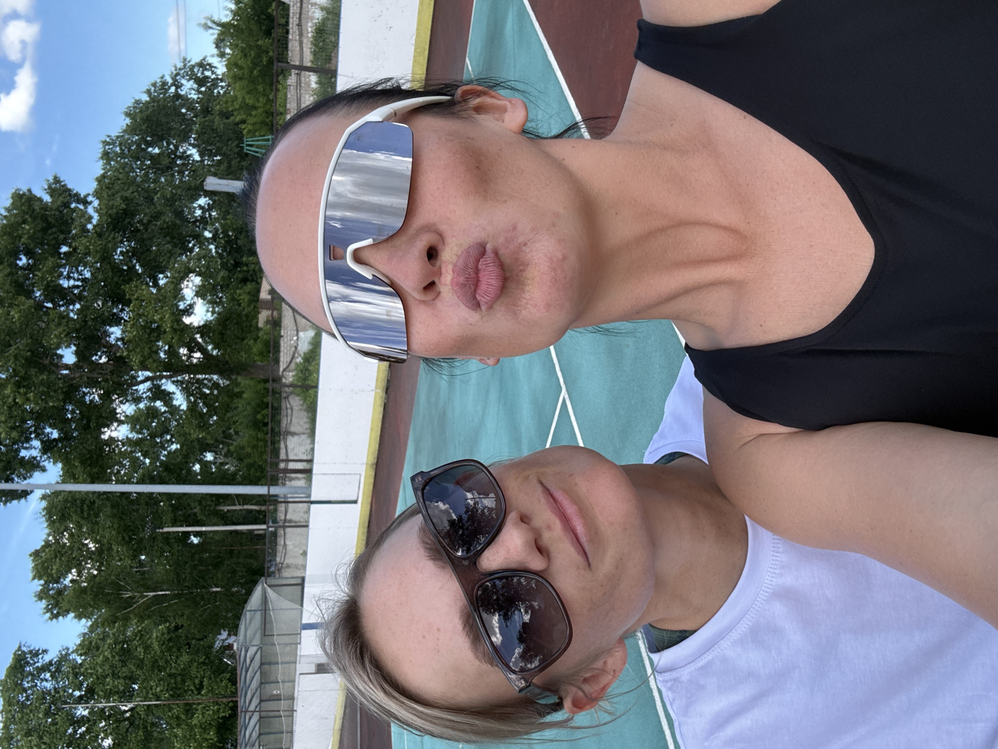 Two women, likely adult females, are captured in a selfie taken outdoors. The woman in the foreground, on the right, is wearing a black tank top and large, mirrored, wrap-around sunglasses, and is puckering her lips as if for a kiss. Her skin appears to have some redness and texture on her cheeks. The woman behind her, on the left, wears a white t-shirt and dark, rectangular sunglasses that reflect the sky and trees. Her expression is relaxed, with a slight smile.
The background suggests an outdoor sports facility. A turquoise and red court, possibly a tennis or roller hockey court, is visible. Beyond the court, a light-colored fence with netting runs along the perimeter, and further in the distance, lush green trees are prominent against a bright blue sky with scattered clouds. The overall setting appears to be a public park or recreational area. Based on the context provided by the city name, this scene is located in Chaikovsky, Russia.