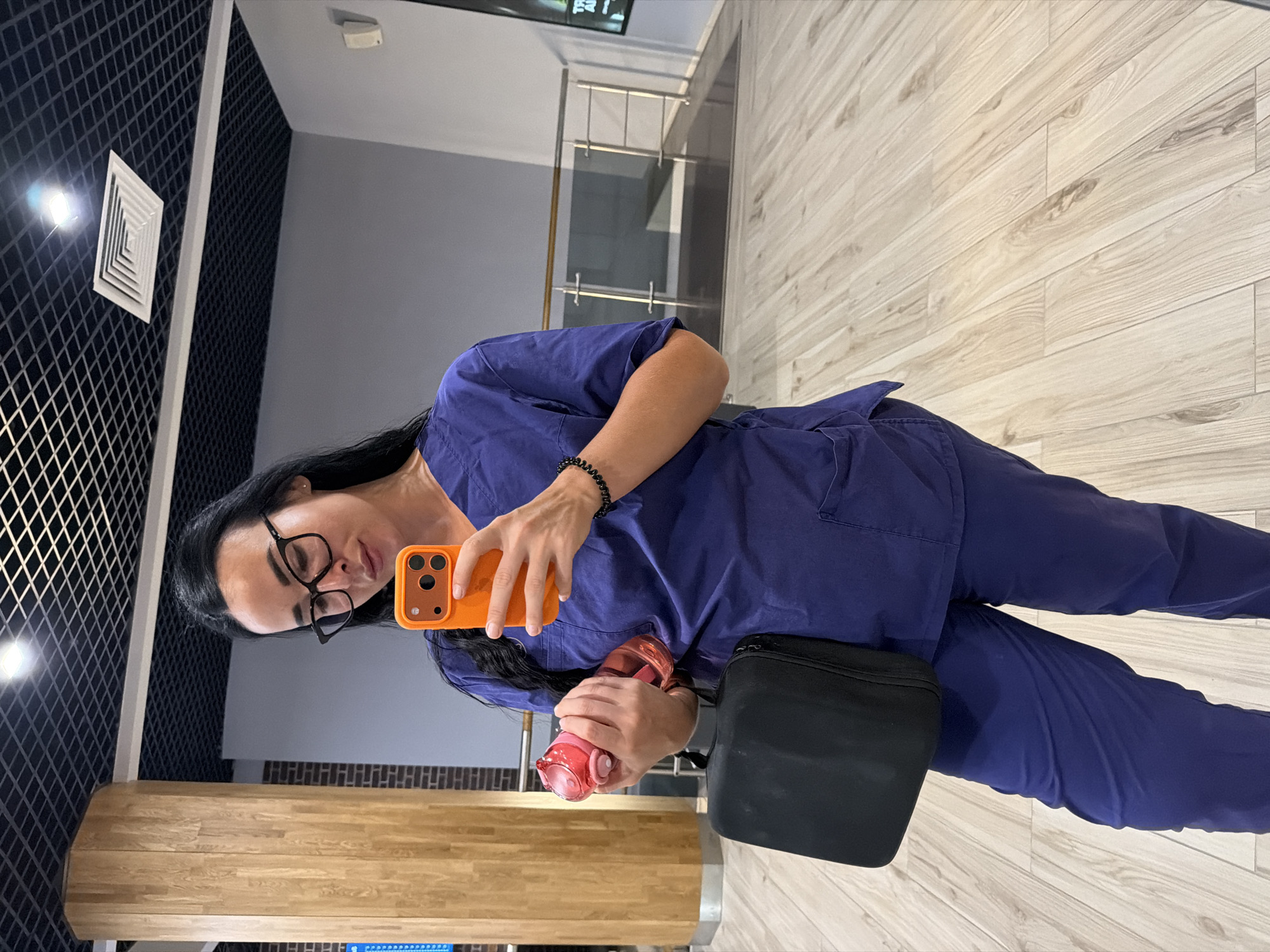 A woman with dark hair, wearing blue scrubs, glasses, and a black bracelet, stands in a well-lit indoor space. She is holding an orange smartphone in her right hand, taking a selfie, and a pink water bottle in her left hand. A black bag hangs from her left arm. The background features a wooden pillar, a modern ceiling with grid panels and lighting, and a grey wall. The floor is made of light-colored wooden planks. The environment appears to be a public indoor space, possibly a medical facility, shopping mall, or office building in Saint Petersburg, Russia.