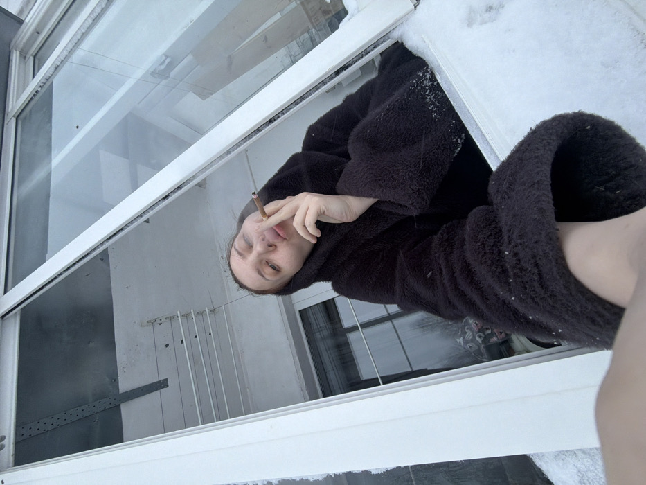 The image captures a person, likely a woman, viewed from outside a window in what appears to be cold, possibly winter, weather. The photograph is rotated, giving a diagonal perspective.
The central subject is a woman with fair skin and dark hair, wearing a dark, possibly black or very dark brown, fuzzy or textured coat. She is positioned within the open window frame, looking directly towards the camera. Her right hand is raised to her mouth, holding a lit cigarette or hand-rolled smoke between her fingers, and a wisp of smoke is visible near her lips, suggesting she has just exhaled. Her expression is somewhat neutral, with a slight hint of a frown or weariness.
The window frame itself is white and appears to be made of PVC or a similar material. There is a layer of snow visible on the exterior sill or frame of the window, particularly noticeable on the upper right side of the frame, reinforcing the impression of cold weather. The view through the window into the interior reveals a muted color palette. Visible elements inside include white walls, reflections of what might be another window or light source, and a metal structure with horizontal bars and vertical lines, possibly a clothes drying rack or a utility shelf, visible in the background through the window.
The perspective is from below, looking up at the person through the window. The overall lighting is soft and diffuse, characteristic of an overcast day or perhaps early morning/late afternoon during winter. No discernable text is visible in the image.