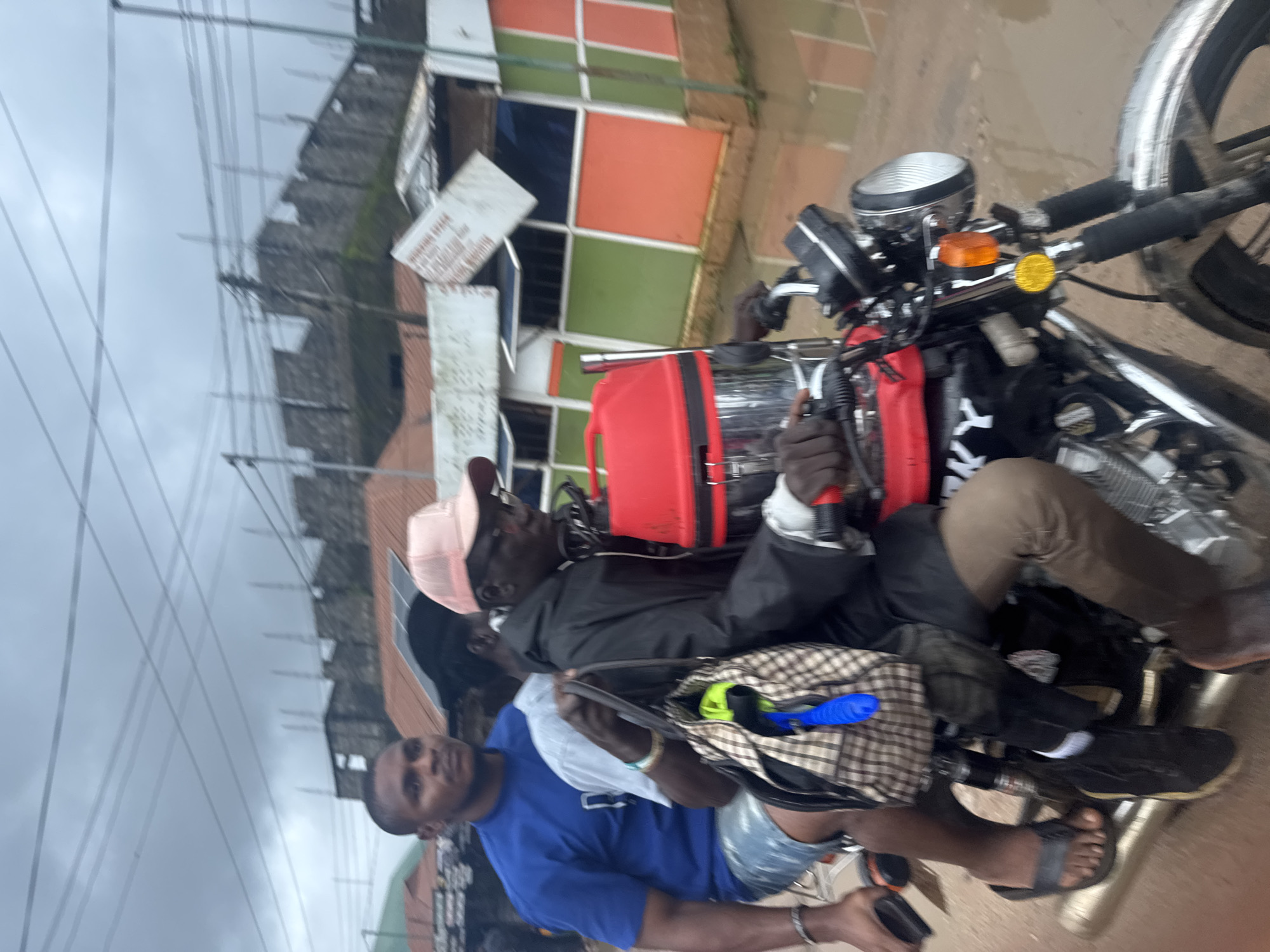 In Ihiagwa, Nigeria, three individuals are riding on a motorcycle. The person in the front, wearing a black jacket and a pink cap, is driving. Behind him, a person in a light blue shirt is seated, and further behind, a person in a blue t-shirt and shorts is the third passenger. The motorcycle is carrying a large red container on its back. The scene appears to be outdoors, with buildings and overhead wires visible in the background. The ground is wet and appears to be muddy, suggesting recent rain or flooding.