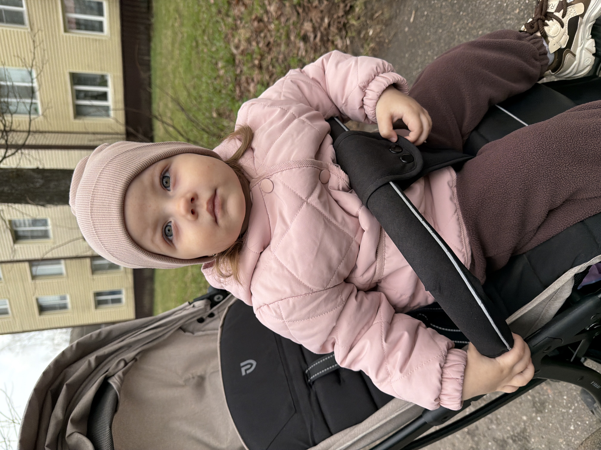 A young child wearing a pink quilted jacket and a matching knit hat is sitting in a stroller. The child also wears brown sweatpants and light-colored sneakers. The stroller is gray and black. The background shows a yellow building with white-framed windows, green grass, and bare trees. The picture appears to be taken outdoors, possibly in Reshetnikovo, Russia.