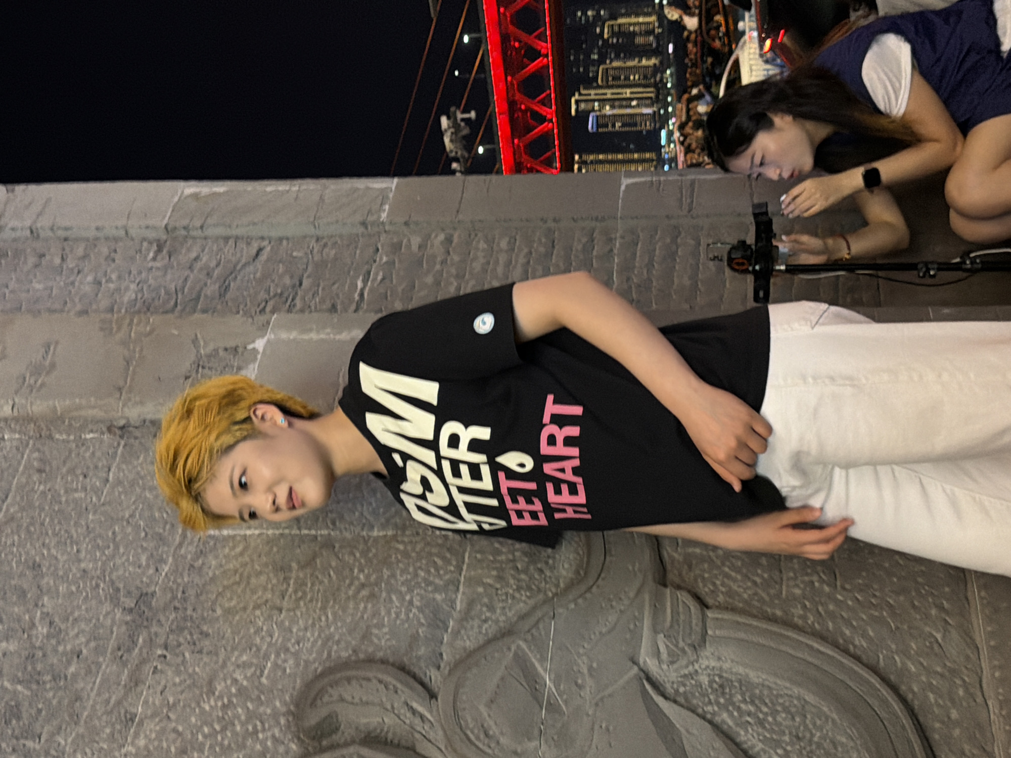 In Yuzhong District, China, a person with short, bright yellow hair stands facing left, leaning against a textured stone wall. They wear a black t-shirt with white and pink lettering that reads "ISM BETTER SWEET HEART" and white pants. Their left hand is resting on their thigh, while their right hand is partially obscured. To the right, a second person kneels and is looking into a camera mounted on a tripod, their hands near their face. In the background, a city skyline with illuminated buildings and a red bridge is visible at night, with blurred figures and vehicles in the foreground.