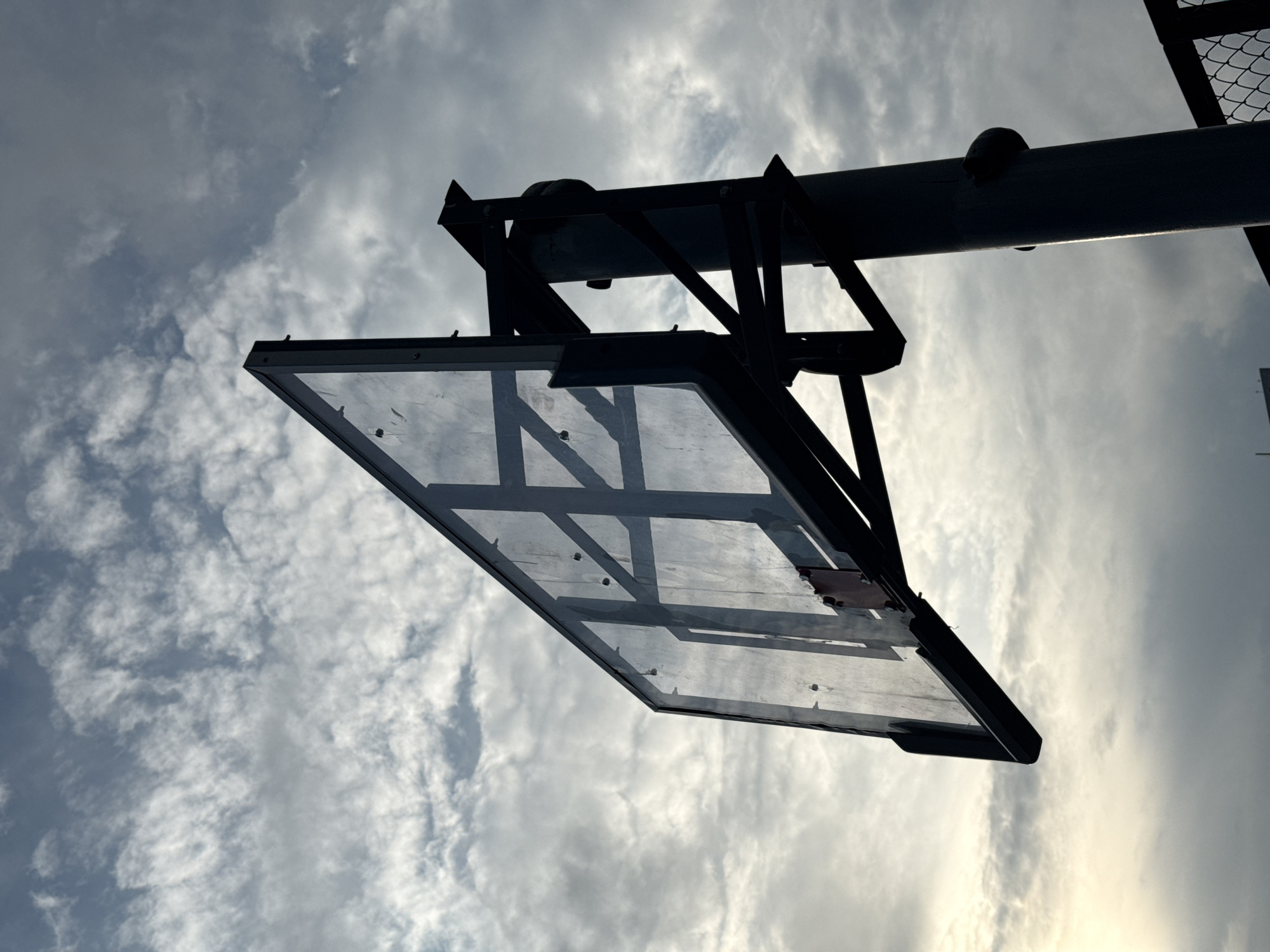 The media file shows a low-angle shot of a basketball backboard and hoop structure against a cloudy sky. The backboard is made of clear material, likely glass or acrylic, with a black metal frame and reinforcement bars visible. The hoop structure is also black metal, with a thick pole extending upwards and to the right, supporting the backboard. A circular, dark object, possibly a small ball or a cap, is visible on the pole. The sky is filled with dramatic, textured clouds, suggesting it might be overcast or that the sun is behind the clouds, creating a diffused light. The overall mood is somewhat dramatic and stark due to the silhouette of the structure against the sky. There is no visible text in the image. The time of day is difficult to pinpoint precisely but the lighting suggests it could be late afternoon or early morning, when the sun is lower. The location is not identifiable from the visual cues.
