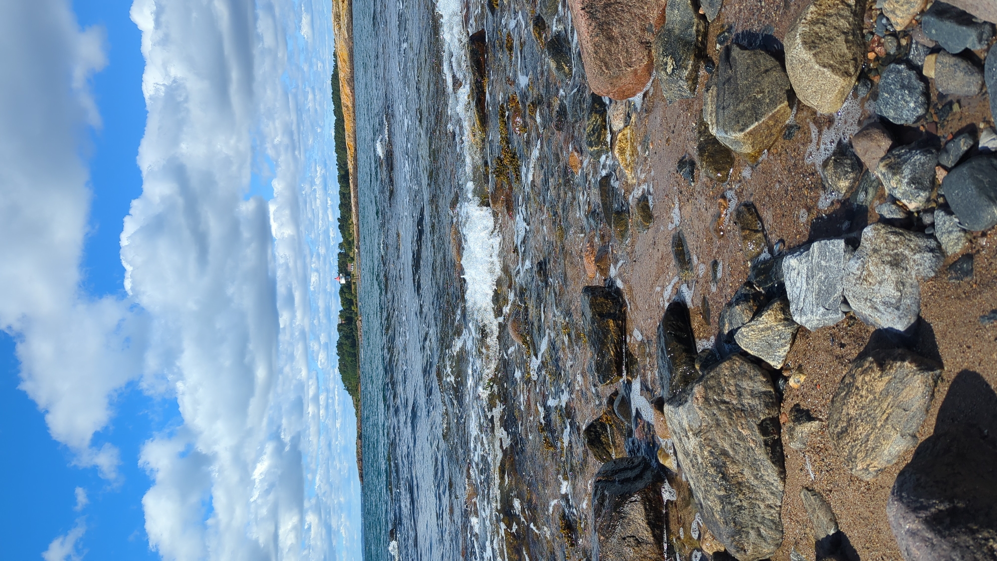 The image depicts a rocky shoreline with gentle waves lapping at the shore. The foreground is filled with various sizes of rocks, some wet from the tide, interspersed with sand and small pebbles. In the middle ground, the sea is visible with a small wave breaking. The background shows a distant landmass covered with green trees, and a white building with a red roof, likely a lighthouse, is situated on a rocky outcrop. The sky above is blue with fluffy white clouds. This scene is consistent with the coastal environment found in Larkollen, Norway.