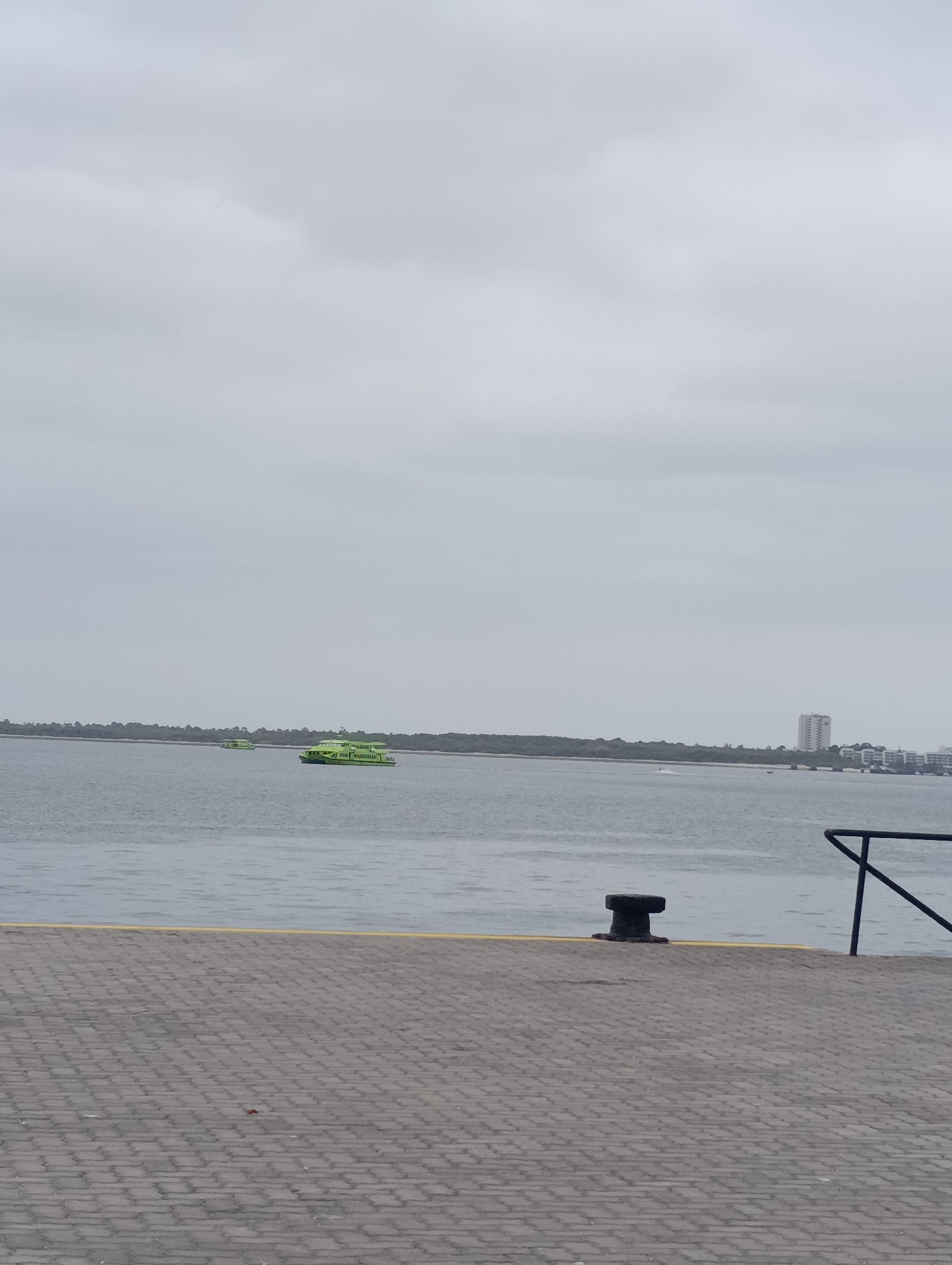 The image captures a waterfront scene in Setúbal, Portugal, under an overcast sky. In the foreground, a paved walkway made of grey interlocking bricks leads towards the water, with a yellow line demarcating the edge. A dark, T-shaped bollard stands on the walkway near the water's edge. To the right, a black metal railing is visible, extending diagonally into the frame.
The water in the mid-ground is calm with ripples. Two bright green boats are traveling on the water. The larger boat in the foreground has text on its side, though it is illegible. The boat behind it is smaller and further away.
In the background, a long, tree-lined strip of land stretches across the horizon. Beyond the land, a tall, light-colored building, likely a high-rise apartment or hotel, stands prominently, with other buildings of varying sizes visible in the distance, suggesting an urban or semi-urban area. The overall atmosphere is subdued due to the cloudy weather.