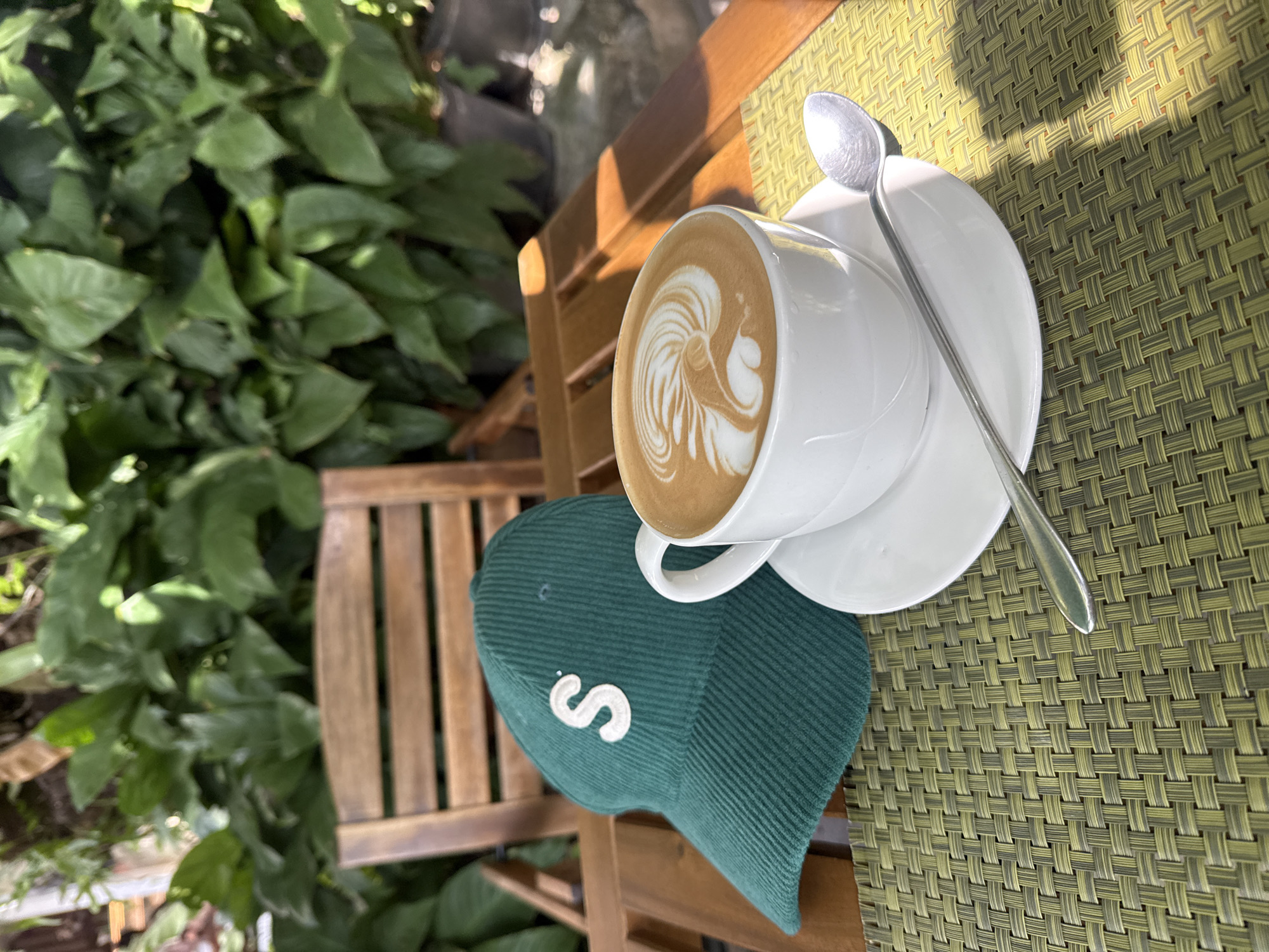 A close-up shot shows a coffee cup with latte art and a green corduroy cap with an "S" logo on a wooden table with a woven placemat. The background is blurred, featuring lush green foliage and a wooden chair, suggesting an outdoor setting in a tropical location like Ko Samui District, Thailand. The sunlight casts shadows on the table, indicating it is daytime.