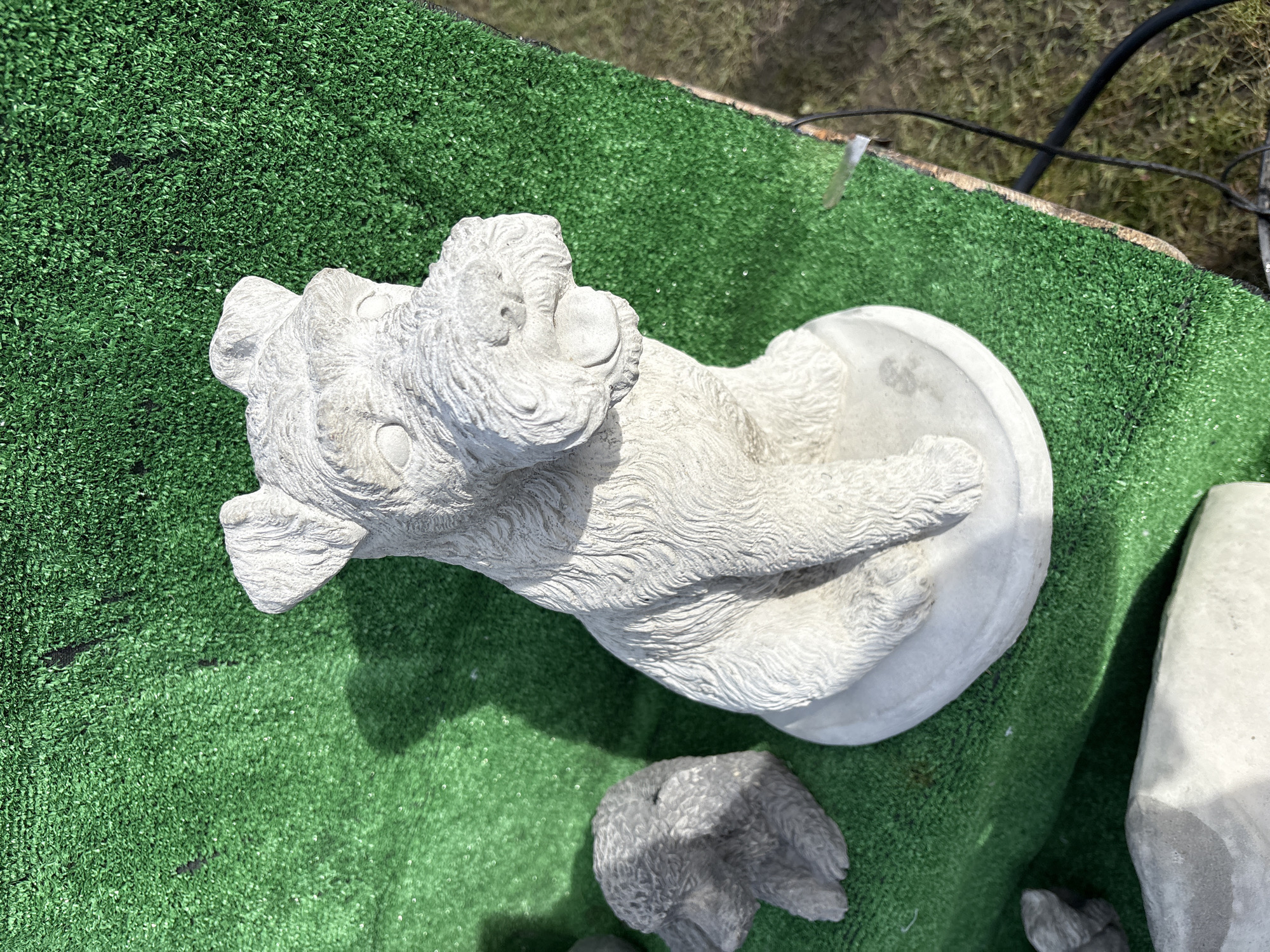 The image depicts a light-colored, detailed concrete statue of a dog lying on its back, paws extended. The statue rests on a patch of artificial green turf. To the lower right of the statue and scattered around it are similarly-colored concrete objects. The surrounding environment, including the city of Chirk, United Kingdom, is not discernible from the image.