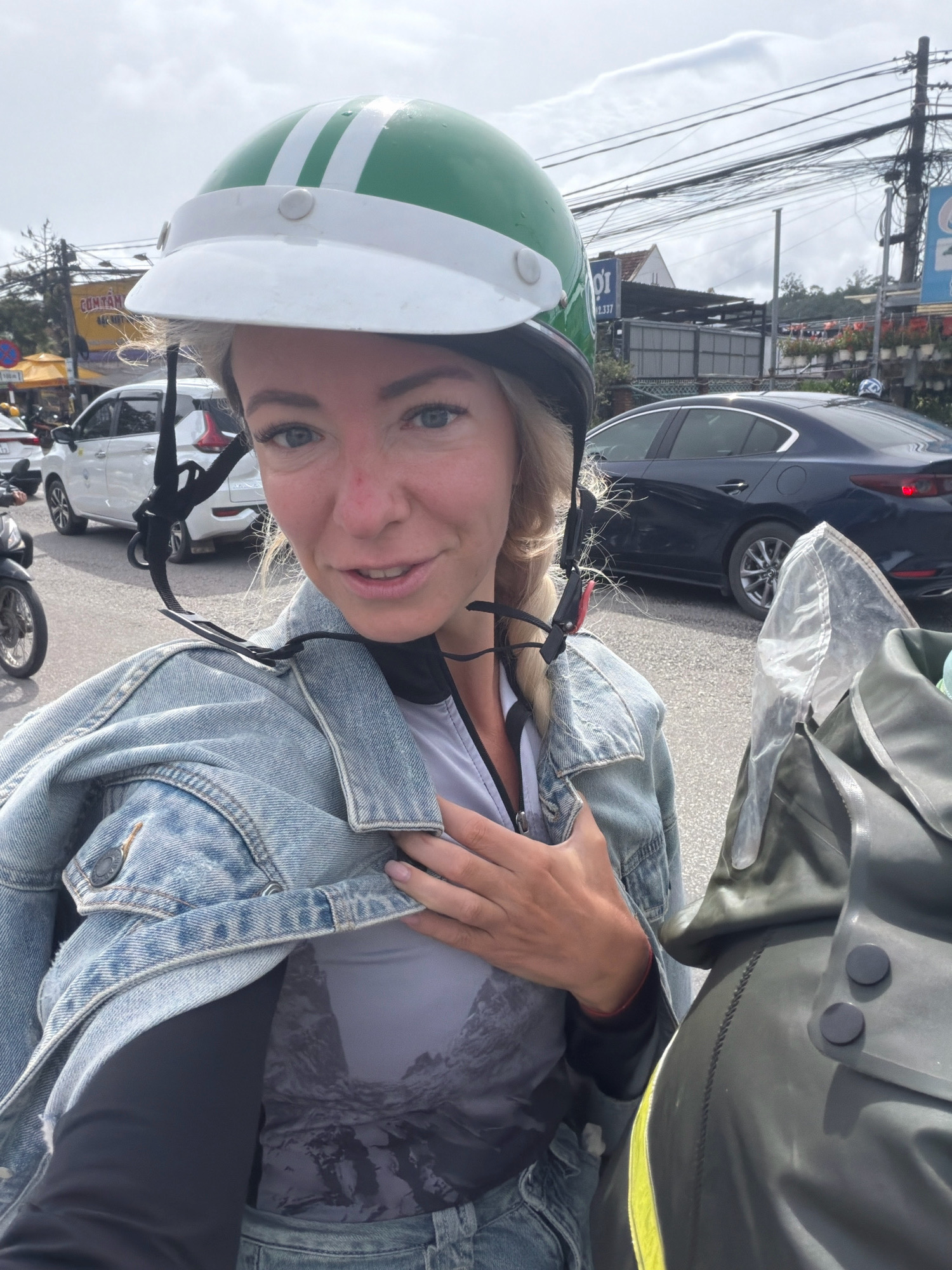 The image is a selfie taken by a blonde woman wearing a green and white helmet and a denim jacket over a gray shirt with a mountain print. She is on a motorcycle in an urban environment. In the background, there are cars, motorcycles, buildings with signs in Vietnamese, and power lines. The location appears to be Lam Vien - Da Lat, Vietnam, as indicated by the distinctive architecture and the presence of a large, modern structure, possibly the city's cultural center, in the background. The sky is overcast. The woman is looking at the camera and smiling slightly. The image is cropped to focus on the woman and her immediate surroundings.
