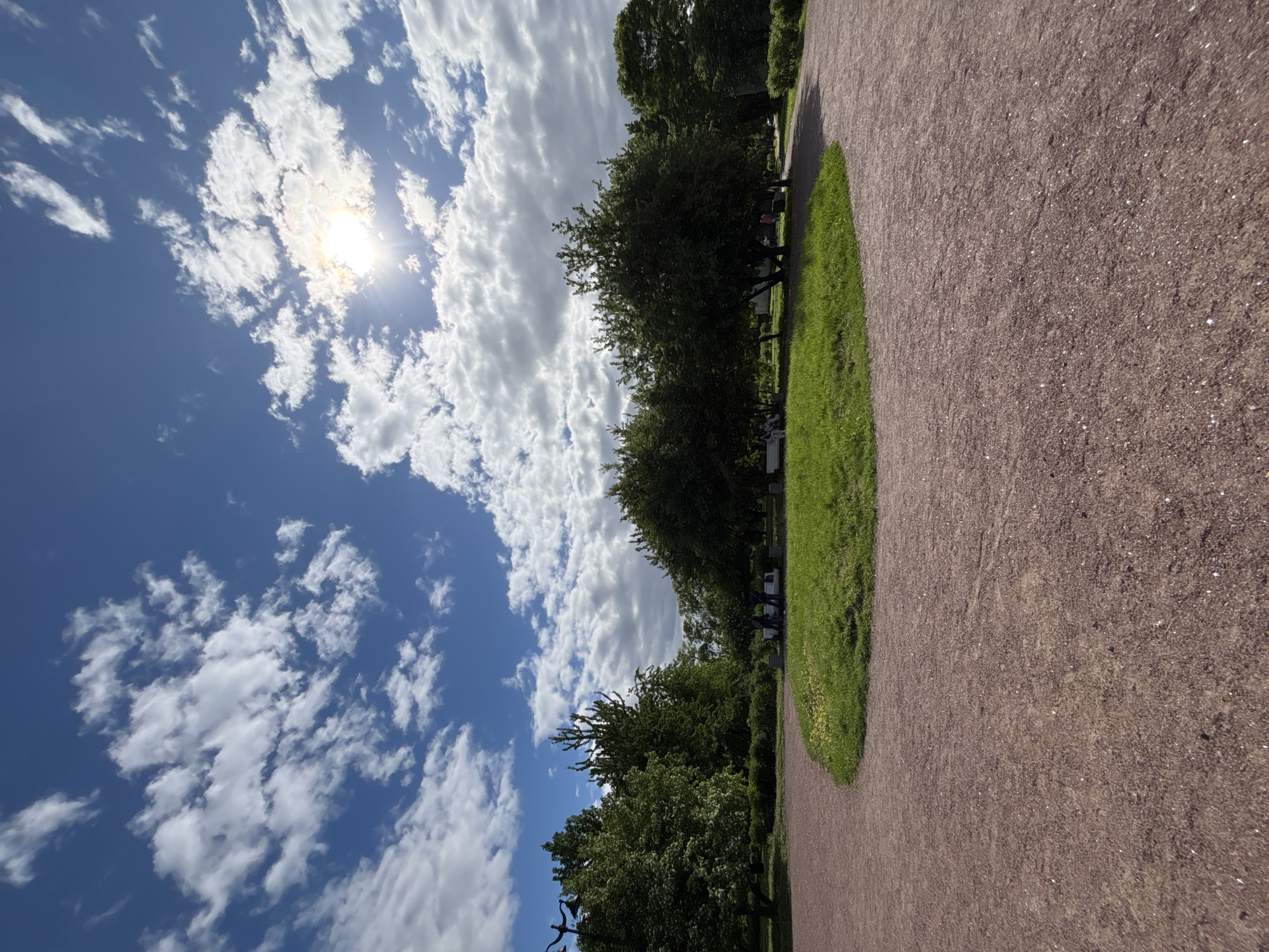 The image shows a scene in Saint Petersburg, Russia, featuring a dirt path running along the right side of the frame. Adjacent to the path is a strip of green grass and trees lining the middle ground. In the background, a bright blue sky is filled with white, fluffy clouds and a sun visible through some clouds. The scene appears to be taken during daylight hours.