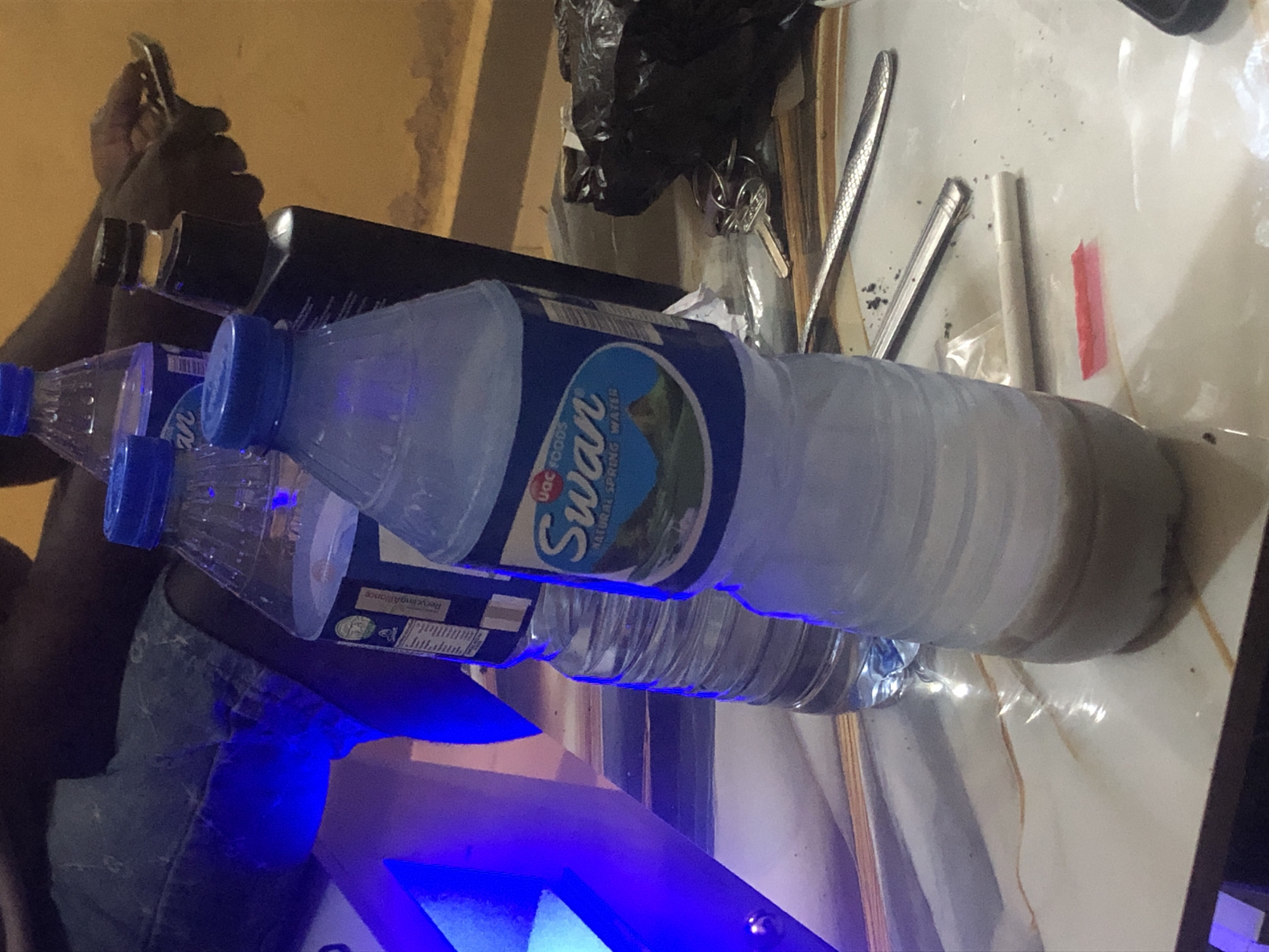 The image depicts a close-up of a table surface with several objects. In the foreground, a partially filled plastic bottle of "Swan Natural Spring Water" is visible, with a light brown liquid at the bottom. To its left, another plastic bottle, also partially filled, with a blue cap is seen. Behind these, a dark-colored bottle with a black cap stands. To the right of the bottles, a black plastic bag contains a set of keys. There are also two metal utensils on the table, one partially visible and the other clearly showing a decorative pattern. A single cigarette and a small piece of red tape are also on the surface. In the background, a person wearing blue denim shorts is seated, with a blue light emanating from the left side of the frame. The wall in the background is a textured orange-yellow. The setting appears to be indoors, with the overall lighting suggesting a dimly lit environment enhanced by artificial light. The presence of a phone and a person's hand in the background suggests recent activity. The scene is consistent with a casual gathering or rest area in Kabong, Nigeria.