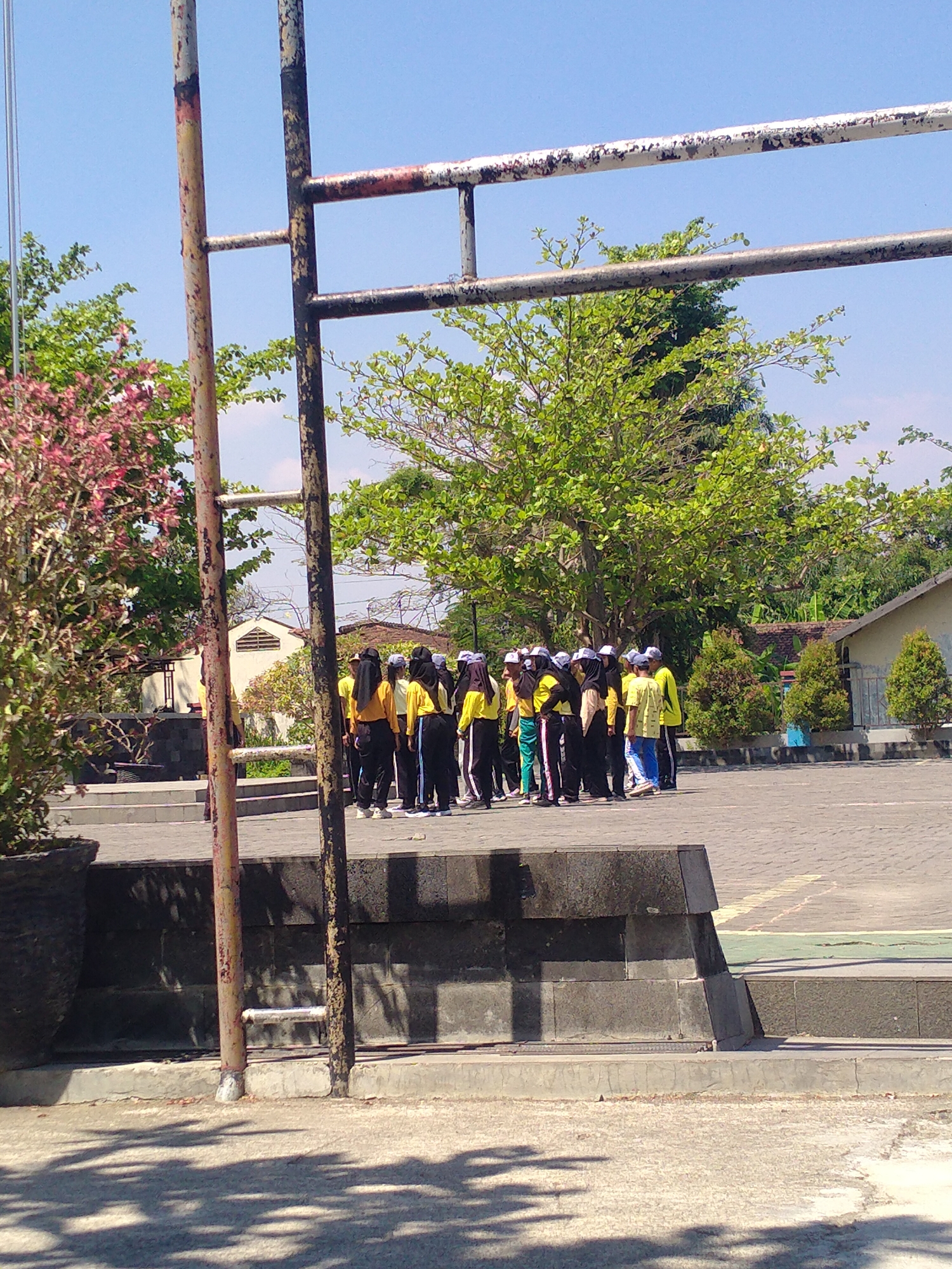 In Begajah, Indonesia, a group of young people, likely students wearing yellow shirts and black pants or skirts, stand in a huddle on a paved outdoor area. Some individuals wear white caps, and a few have purple head coverings. The group appears to be assembled for an activity, possibly physical education or a school event. They are standing in front of a raised, tiered stone structure. In the foreground, a large plant in a dark pot sits to the left, and shadows from trees and a metal structure are cast on the ground. A rusty metal pole, part of what appears to be a goalpost or sports equipment, stands prominently in the left-center of the frame. Behind the group, trees with dense green foliage and a building with a white facade and a tiled roof are visible under a clear blue sky. The scene suggests a sunny day in an outdoor school or recreational area.