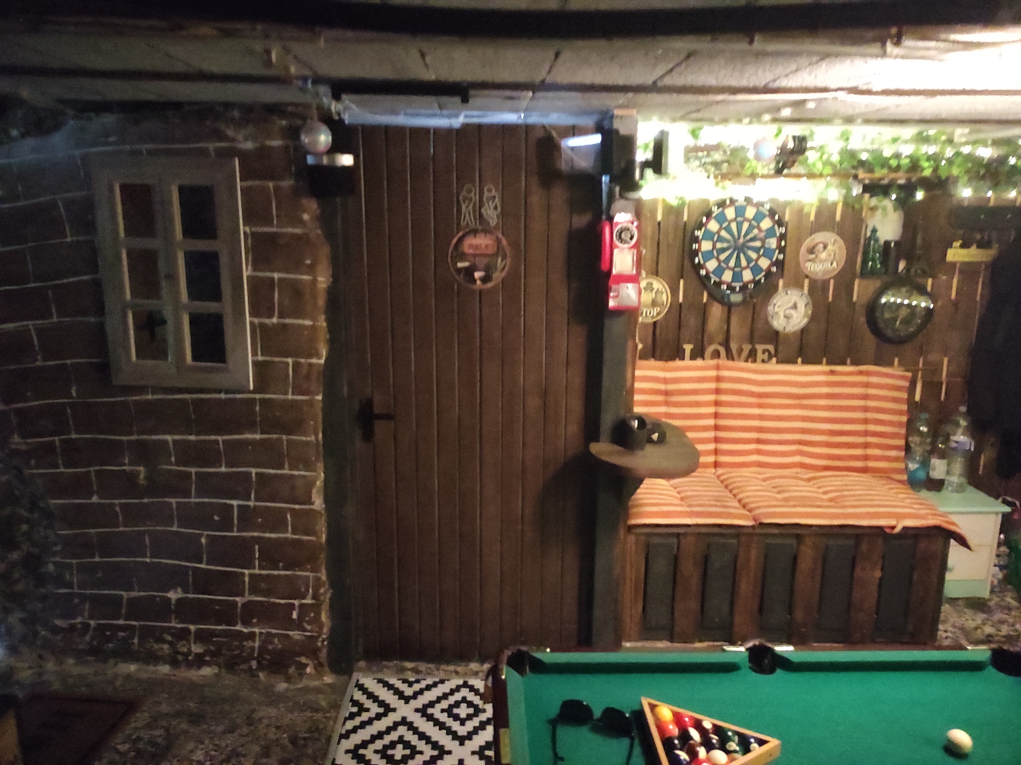 The image depicts a dimly lit, rustic room that appears to be a bar or entertainment area, potentially a basement or a converted garage. The walls are primarily constructed of dark brick on the left and wooden planks on the right. In the center of the image is a dark wooden door with a vertical plank design. Above the door, a sign with two stylized figures resembling a man and a woman, along with the word "TOILET" is visible. To the right of the door, a red vintage-style gas pump is mounted on the wall.
Further to the right, a rustic wooden wall is adorned with several decorative items, including a dartboard, circular plaques with text and imagery such as "TEQUILA," and a round mirror. Below these decorations, the word "LOVE" is spelled out in large, rustic letters. A bench with a striped cushion (alternating orange and white) is positioned in front of this wall.
On the far left of the image, a small, multi-paned window is set into the brick wall. A disco ball hangs from the ceiling near the window. In the foreground, a pool table with a green felt surface is visible, with a rack of colorful billiard balls and a single white ball on the table. A black and white geometric patterned rug is placed on the floor in front of the pool table.
The overall impression is of a casual, leisure-oriented space with a distinct pub or man-cave aesthetic. The lighting is low, with some string lights visible in the upper right portion of the image, suggesting an evening or nighttime setting. There are no people visible in the image. The location is identified as Waldbronn, Germany.