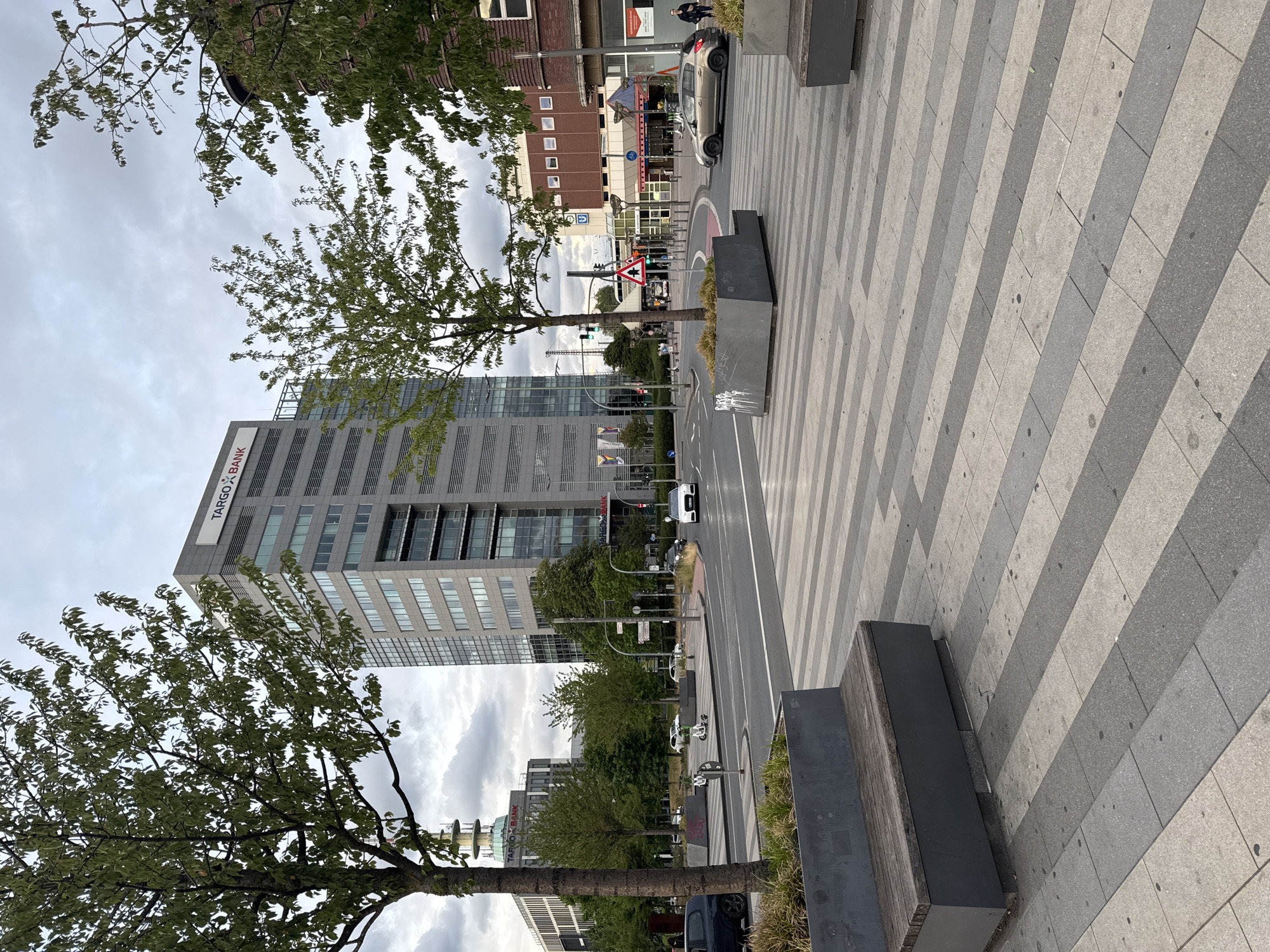 The image depicts a street scene in Duisburg, Germany, specifically near the TARGOBANK building. In the foreground, a modern streetscape features a paved walkway with a repeating pattern of light and dark grey rectangular tiles. Two dark grey planters, each with a wooden bench, are positioned on the tiled area. One planter in the foreground contains dry grass. Beyond this, a multi-lane road is visible, with a white sedan driving in the right lane and a light-colored sedan parked in the right lane further down the road. Trees with green foliage line the street.
Dominating the background is a tall, modern office building with a grey facade and numerous windows, bearing the TARGOBANK logo at the top. To the left of this building, another, shorter building with a dome-like structure and the TARGOBANK logo is visible. Further in the distance, more buildings and streetlights are present under an overcast sky. Traffic signals are visible, indicating an intersection. A few individuals are discernible on the sidewalks and near the buildings, including a person walking a dog on the far right and a person standing near a scooter on the left. The overall impression is that of an urban environment during the daytime.