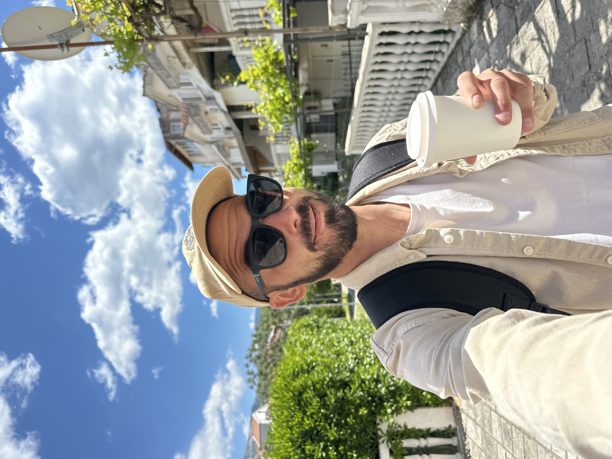 The image is a selfie taken in Budva, Montenegro. A man with a beard, wearing sunglasses and a hat, holds a white cup. He's wearing a white t-shirt and a light beige button-down shirt, with a black strap across his chest. Buildings are visible in the background, with green vines and foliage. The sky is blue with white clouds. The ground appears to be a stone walkway or sidewalk. There's a satellite dish mounted on a building in the background.