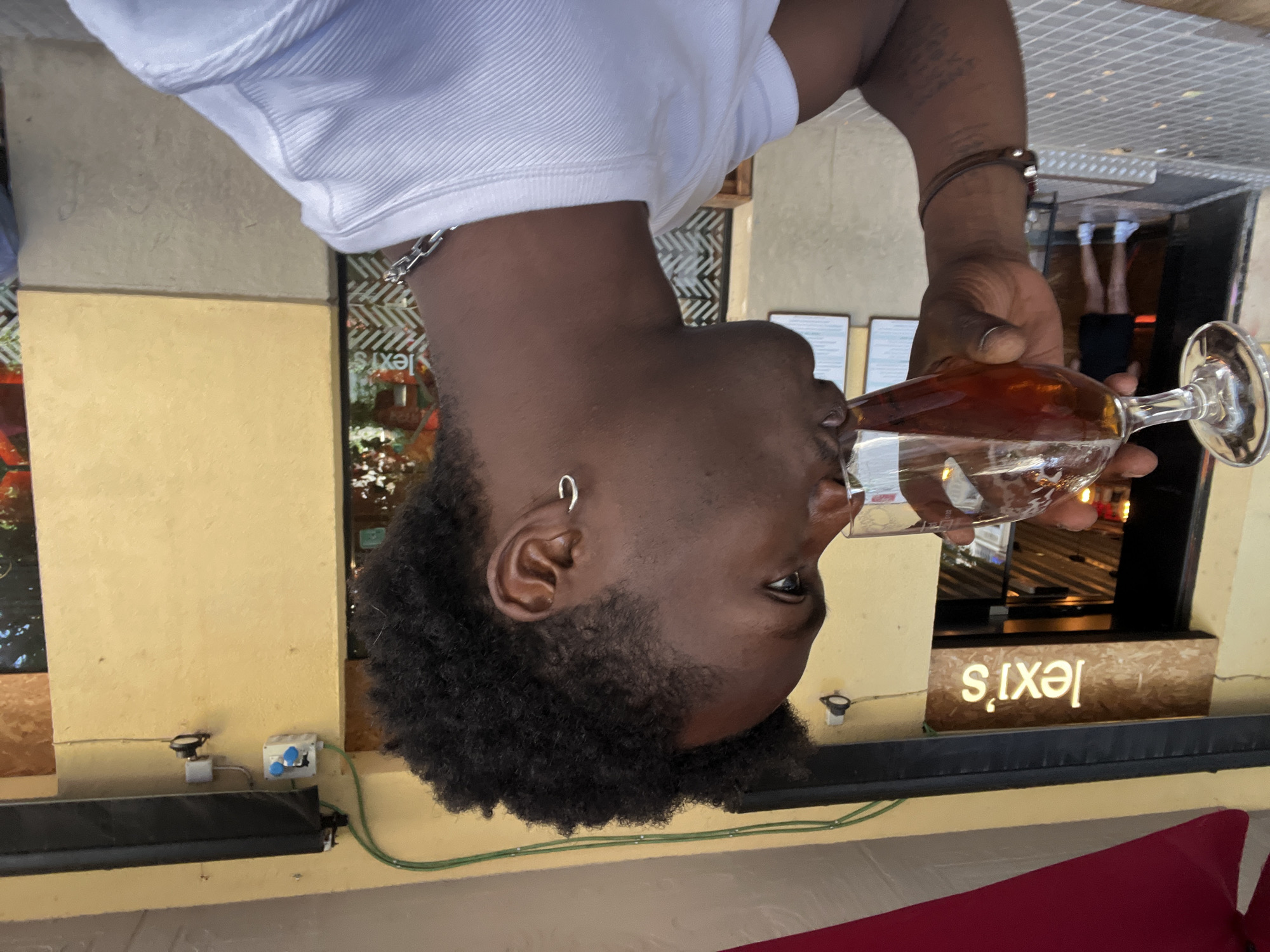 In Barcelona, Spain, a young Black man with dark, curly hair and an earring is captured in a medium close-up shot, drinking from a stemmed glass of amber-colored liquid, likely beer. He is wearing a white t-shirt and a silver chain necklace. The background reveals the exterior of a building with a yellow facade. A sign above an entrance reads "lexi's" in illuminated white letters against a wood-grained background. The entrance appears to be a restaurant or bar. Another person, a woman wearing a black dress and white sneakers, is visible through the doorway in the background. The ground is paved with patterned tiles, and a red awning extends over the building. Electrical conduits and fixtures are visible on the building's exterior, suggesting an outdoor seating area.
