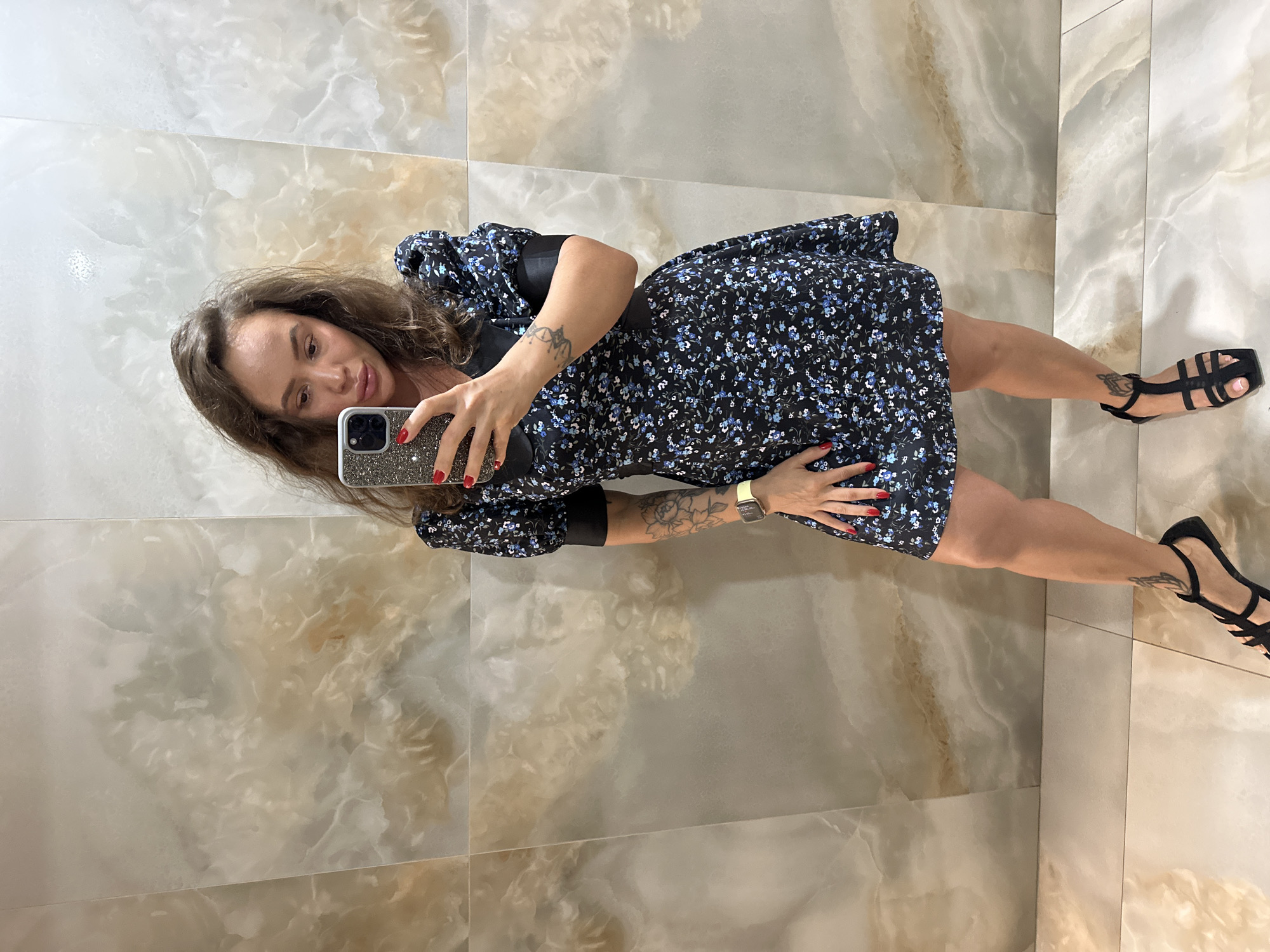 In Duvankul', Russia, a woman stands in front of a wall tiled with marble-like slabs, taking a selfie with her smartphone. She is wearing a short, black, floral-print dress with puffy sleeves and black trim. Her left arm, visible in the shot, is adorned with a tattoo of a flower. A smartwatch with a yellow band is visible on her left wrist. She is also wearing black, strappy sandals. The tiled floor is also visible.