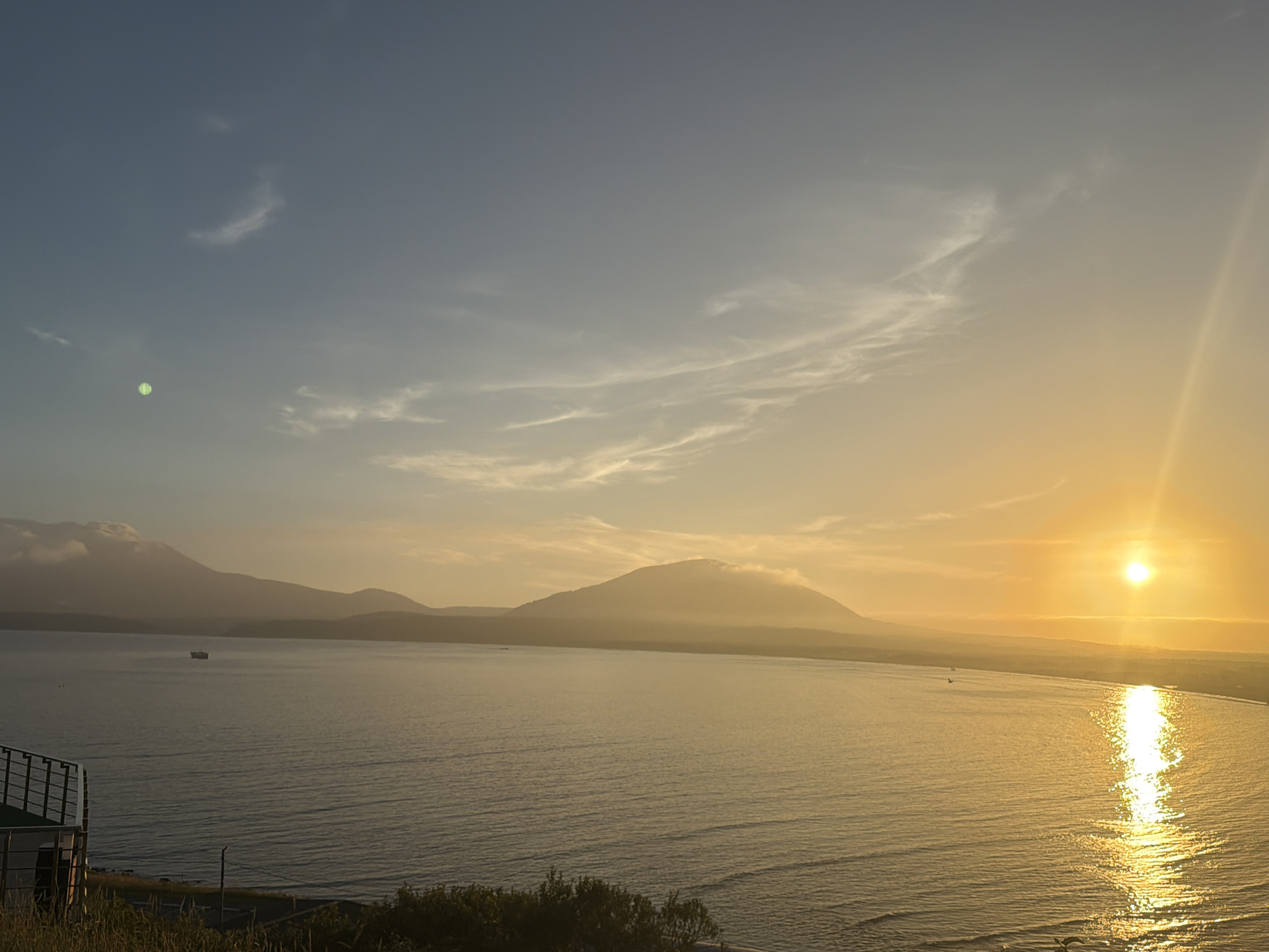 The image depicts a tranquil sunrise over a body of water, likely a bay or sea, with distant mountains forming the horizon. The sky transitions from a pale blue at the top to a warm golden hue near the horizon, illuminated by a bright, low-hanging sun. Wispy clouds are scattered across the sky, catching the sunlight.
In the foreground, a railing and some grassy vegetation suggest an elevated viewpoint, possibly a balcony or a hill overlooking the scene. The water is calm, reflecting the golden light of the sunrise. A small ship is visible in the middle distance, cruising on the water. The shoreline in the far distance is lined with what appears to be a beach and possibly some coastal vegetation or low buildings.
The overall atmosphere is peaceful and serene, characteristic of a sunrise over a coastal landscape. This scene is consistent with the natural beauty found in and around Yuzhno-Kurilsk, Russia, a city situated on Kunashir Island in the Kuril Islands, known for its coastal scenery and volcanic landscapes.