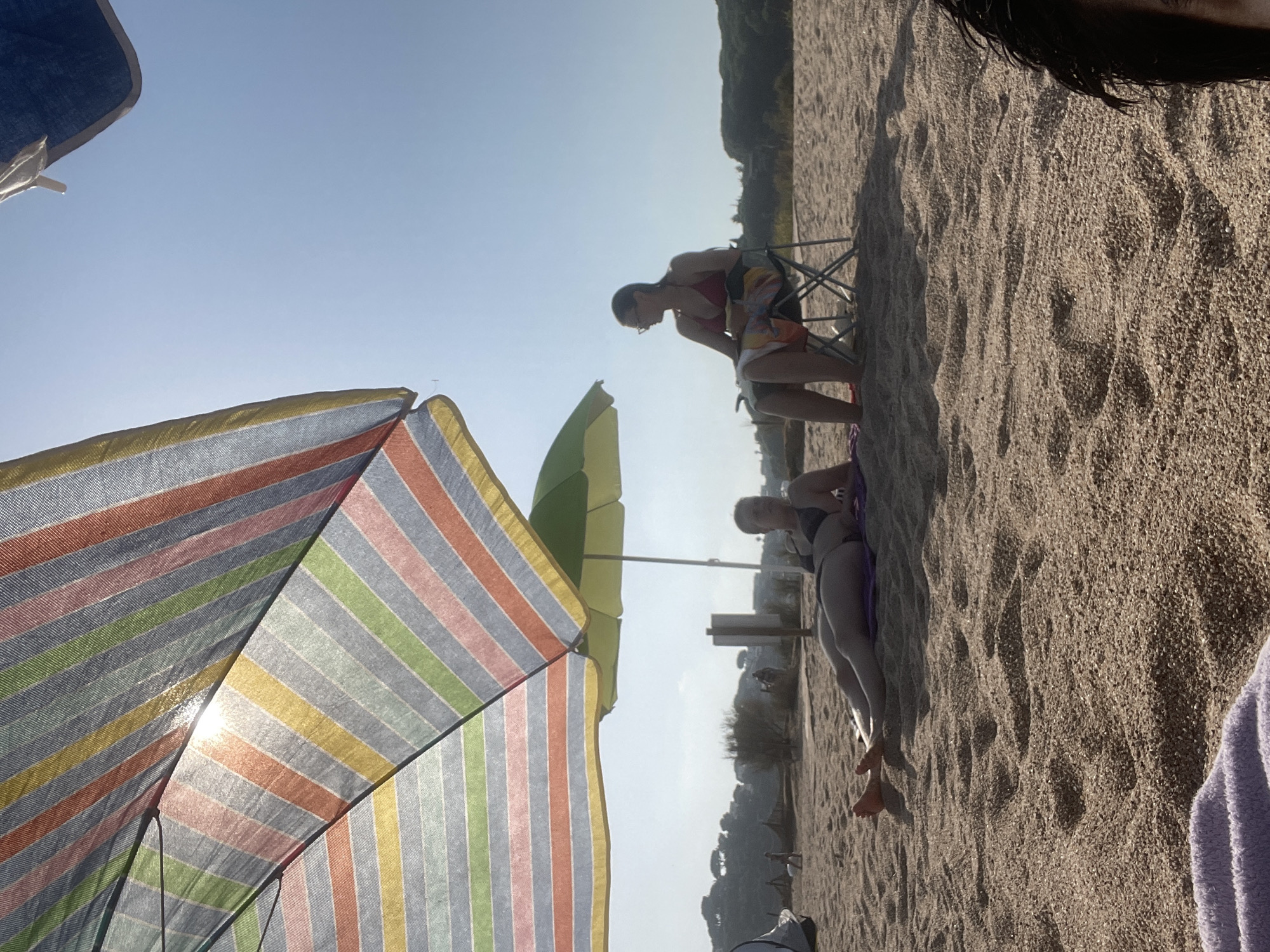 The image depicts a beach scene in Arenys de Mar, Spain. Two women are present. One woman is lying on her stomach on a purple towel on the sand, wearing a dark bikini. The other woman is sitting in a folding chair under a beach umbrella, wearing a red bikini and a patterned towel draped over her legs. She is looking to the left. A large, colorful striped beach umbrella is prominently featured in the foreground, partially obscuring the view of the sky and the sun. A green umbrella is visible in the background. The beach is covered in light-colored sand and small pebbles. In the distance, there are some trees and hills, suggesting a coastal landscape. The sky is clear and bright blue.