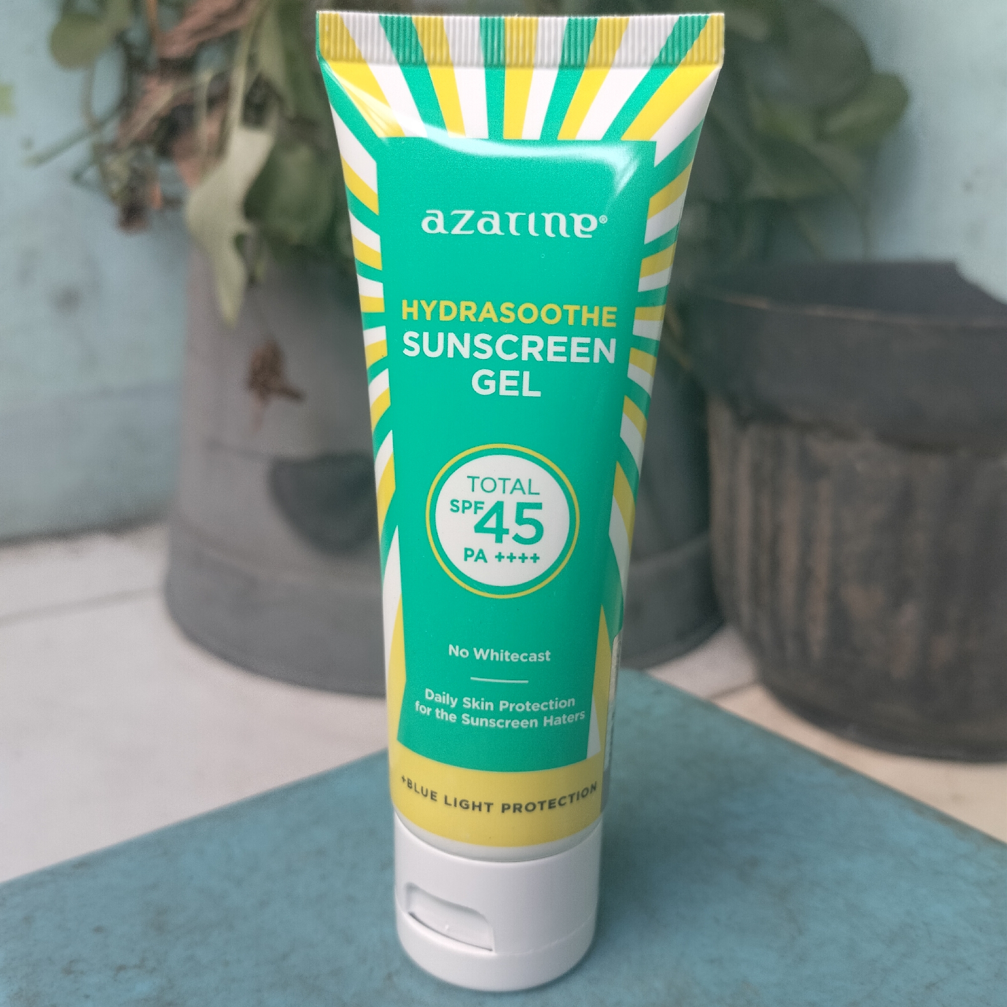 An Azarine Hydrasoothe Sunscreen Gel tube is displayed prominently in the foreground, set against a softly blurred background. The tube, with its distinctive teal and yellow striped design, is oriented vertically and slightly angled. The text on the tube is clear and legible, indicating "azarine," "HYDRASOOTHE SUNSCREEN GEL," "TOTAL SPF 45," "PA ++++," "No Whitecast," "Daily Skin Protection for the Sunscreen Haters," and "+BLUE LIGHT PROTECTION." The cap of the tube is white and has a small opening. The object rests on a textured blue surface. Behind the tube, the background features out-of-focus elements that suggest an indoor or sheltered outdoor environment, possibly a plant in a pot and another dark, textured object that could be a planter or a piece of furniture. The lighting is even, highlighting the product. The scene appears to be staged for a product photograph. There is no indication of a specific location within Tangerang, Indonesia, based on the visual information provided.