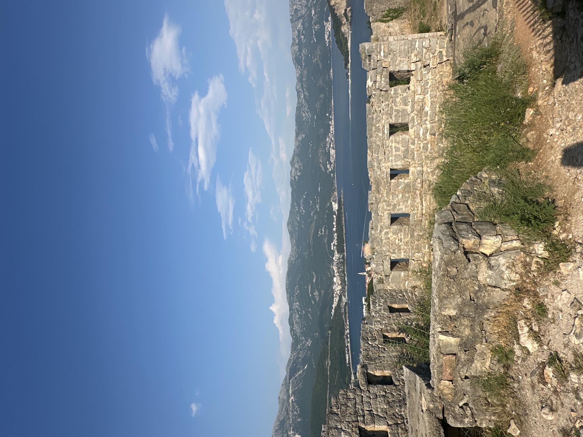 The image shows a view of the Bay of Kotor and the mountains of Montenegro from the ruins of a stone fortress. The fortress walls have small windows or openings along the side. The sky is blue with some clouds. The location is likely within or near the city of Budva, Montenegro, given its proximity to the Bay of Kotor. The fortress structure is made of rough-hewn stone, and there is some vegetation growing on the walls and surrounding area. The scene is taken during daylight with bright sunshine.