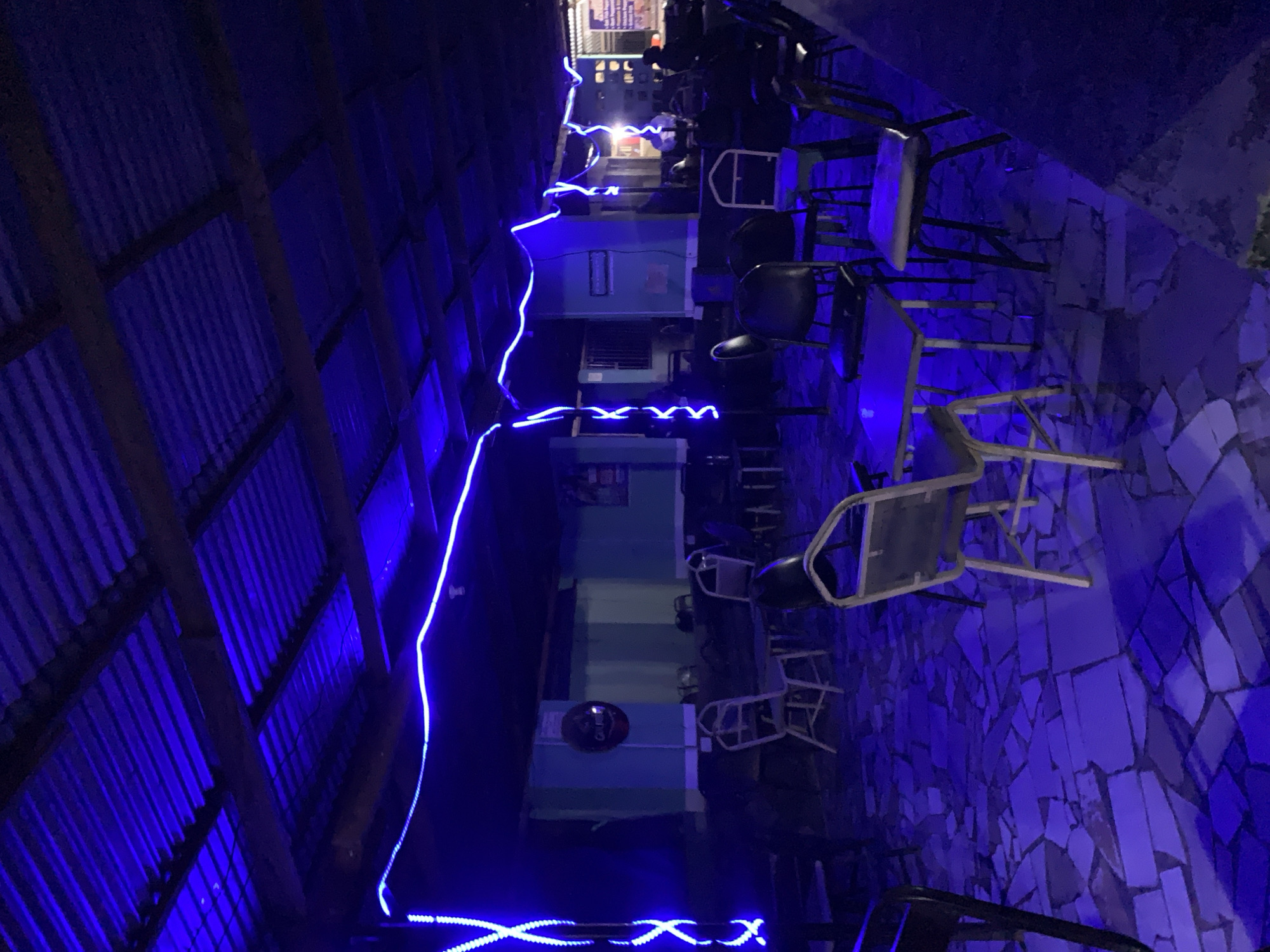The image depicts a dimly lit, semi-enclosed outdoor seating area at night, located in New Stadium, Nigeria. The space is primarily illuminated by blue LED string lights draped across a corrugated metal roof, which is supported by dark wooden beams. The floor is paved with irregular, light-colored flagstones or broken tiles, creating a mosaic pattern. Light blue walls form the background, with a circular "Gulder" brand sign centrally placed on one wall. Numerous metal-framed chairs with upholstered seats and small, light-topped tables are arranged throughout the area. A vertical pole on the left is wrapped with blue LED lights in an 'X' pattern. In the mid-ground and background, a few indistinct figures are faintly discernible, seated at tables. A dark, solid structure, possibly a counter or bar, occupies the right foreground. Additional signage with obscured text is visible on a far background wall.