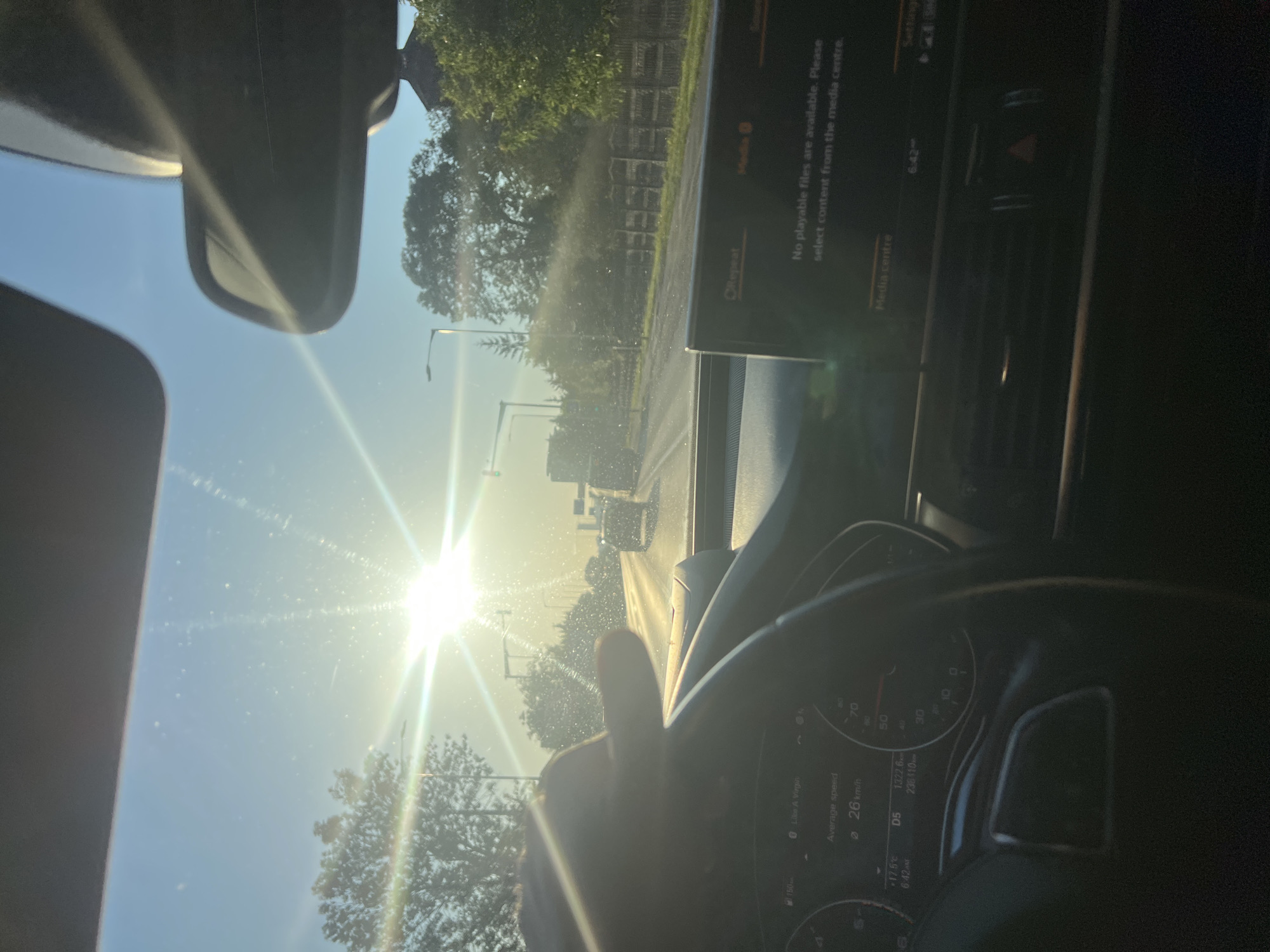 This is a first-person view from inside a car driving on a road. The sun is shining brightly, creating lens flare and glare on the windshield. A hand is visible on the steering wheel. The dashboard displays information including average speed of 26 km/h, an outside temperature of 17.5°C, and the time 6:42 AM. A media screen on the dashboard shows the message "No playable files are available. Please select content from the media centre." The road ahead has other vehicles and trees lining the sides. The environment appears to be a suburban or rural area. The location is identified as Florești, Romania.