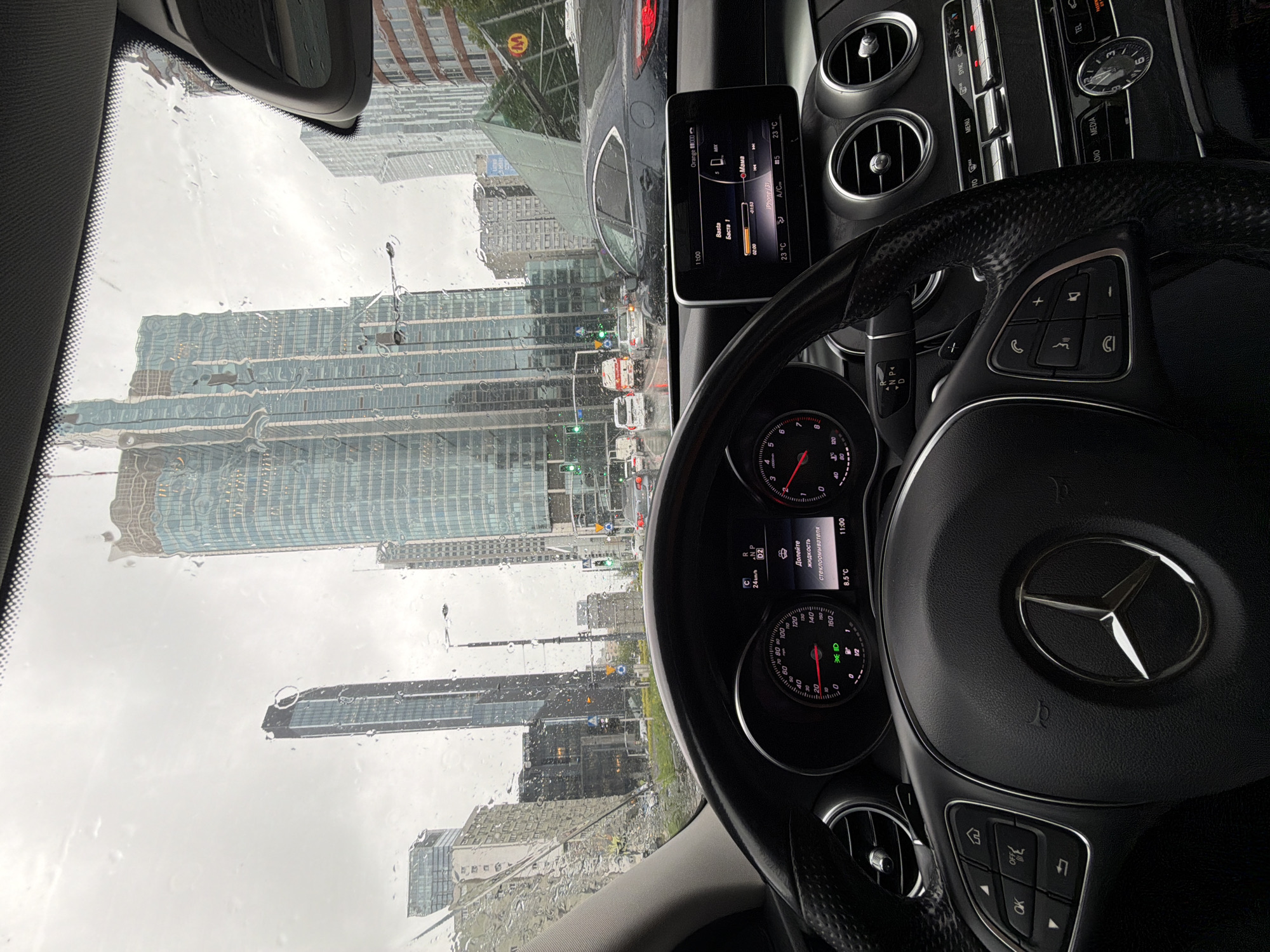 The image, taken from inside a Mercedes-Benz car, shows a cityscape visible through the rain-streaked windshield. The buildings suggest a modern urban setting with tall skyscrapers. Notably, the display screen in the car is in Russian. A yellow "M" sign is visible outside. The dashboard details include a clock set at 11:00 and a temperature of 23°C.