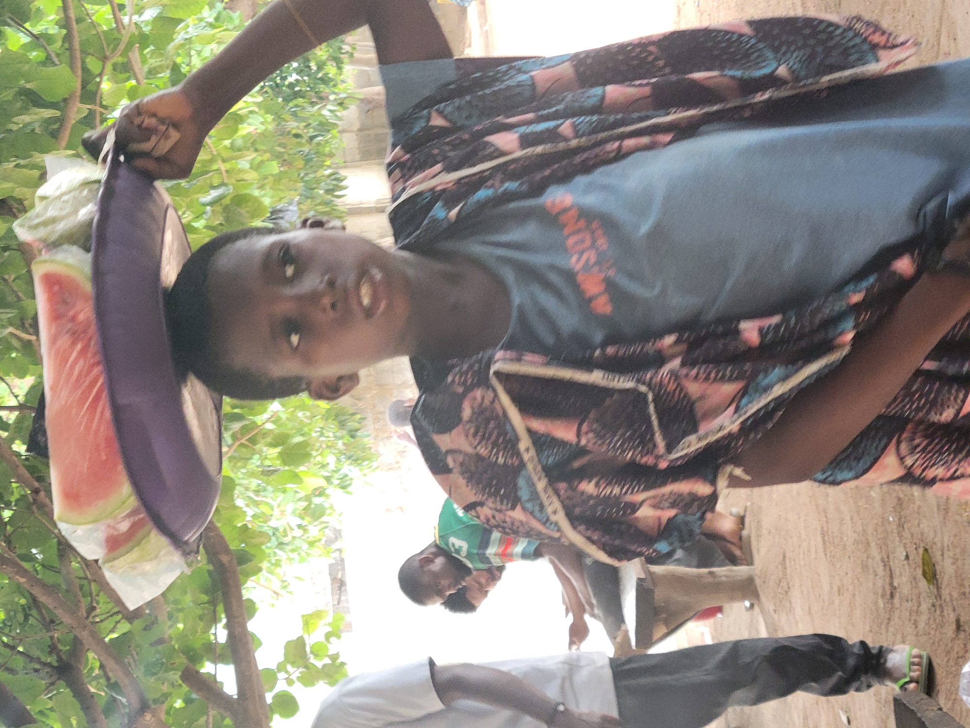 A young person in Madalla, Nigeria, carries watermelon slices in a purple container on their head. They wear a blue shirt under a patterned wrap. Other people are visible in the background, and the setting appears to be outdoors.