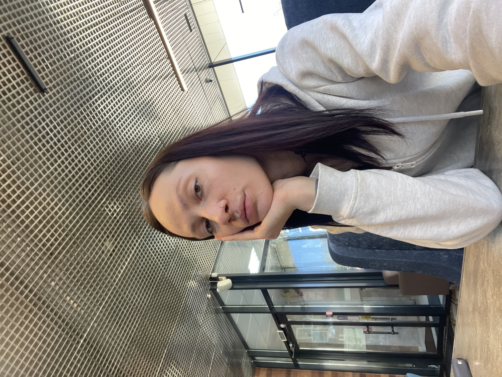 A young woman with long, dark brown hair is resting her face in her hand, looking at the camera. She is wearing a light gray hooded sweatshirt. She is sitting at a table inside a building with a grid-patterned wall. Behind her is a glass-paneled door, and the windows reveal an outdoor view. The location is Naberezhnye Chelny, Russia.
