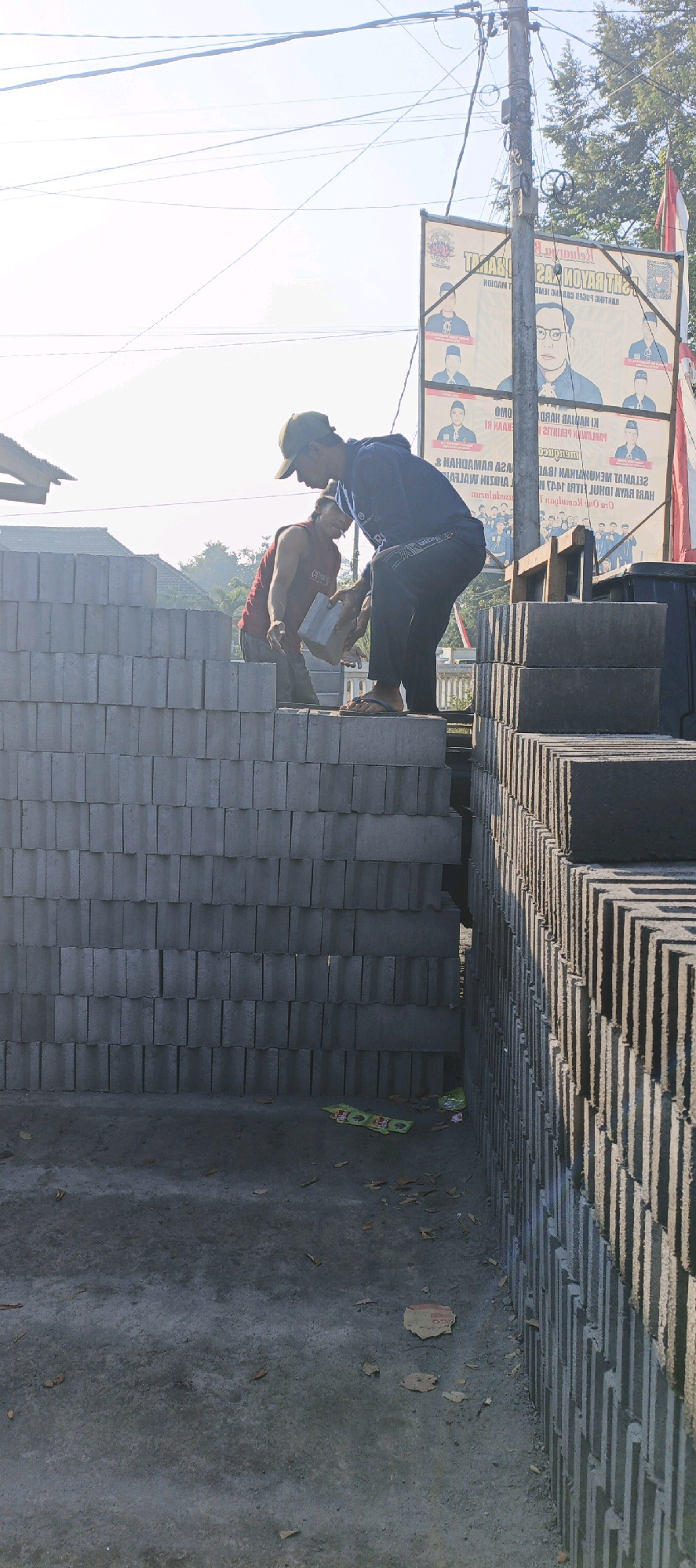 In Kasiyan, Indonesia, two men are working in an outdoor area stacked high with grey concrete blocks. The man on the left, wearing a red tank top and a cap, is reaching for a block. The man on the right, wearing a blue hooded sweatshirt and a cap, is holding a grey block and appears to be placing it onto the stack. The stacked blocks form a wall-like structure. In the background, a large banner with images of men and text in Indonesian is visible, along with power lines and trees. The ground is concrete and scattered with some leaves and small pieces of debris.