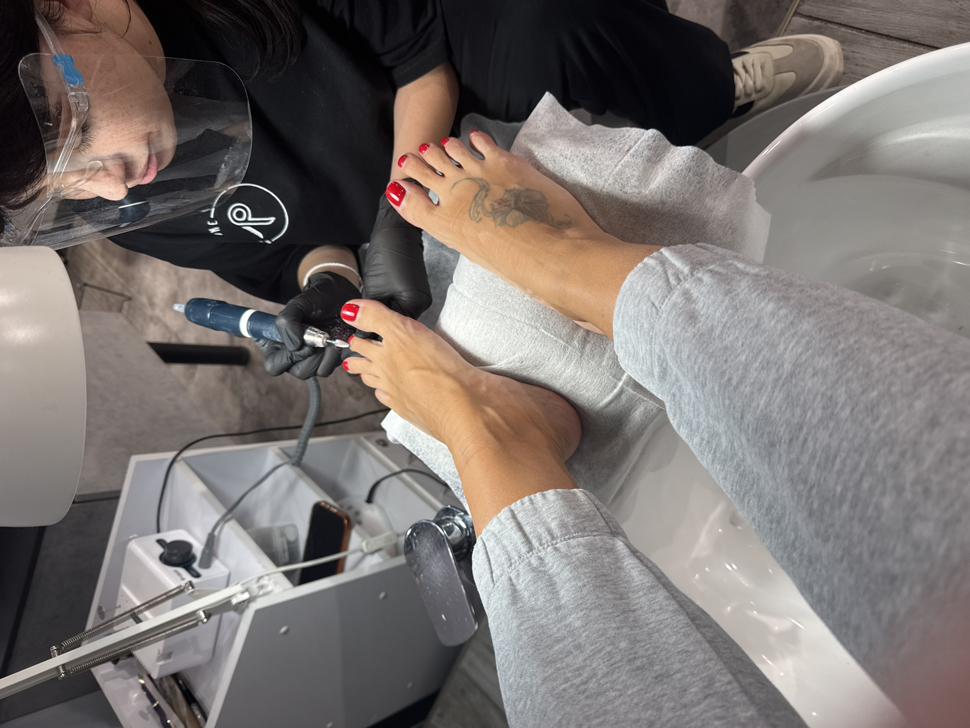A woman wearing a face shield and black gloves is giving a pedicure to a client with red nail polish and a tattoo on their foot. The client is wearing grey sweatpants and sitting next to a white basin filled with water. A white pedicure station is visible with a phone inside. An individual wearing a black shirt and tan sneakers is visible behind the pedicure professional. The location appears to be a nail salon or spa. There is no information to suggest the location is Sochi, Russia.
