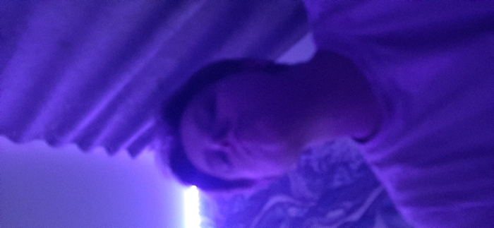 The image captures an indoor scene in the city of Saratov, Russia, under conditions of heavy purple light. A single individual, appearing as a young person, occupies the lower central portion of the frame, seen from the upper torso. The individual's face is directed upwards toward the camera, with the mouth distinctly pursed. They wear a light-colored, short-sleeved garment. The immediate background to the right features a vertically corrugated or folded textured surface, also bathed in purple illumination. In the upper left quadrant, a bright horizontal light source, consistent with an LED strip, is visible, emitting a white-purple glow. The overall scene is characterized by low light quality and blur.