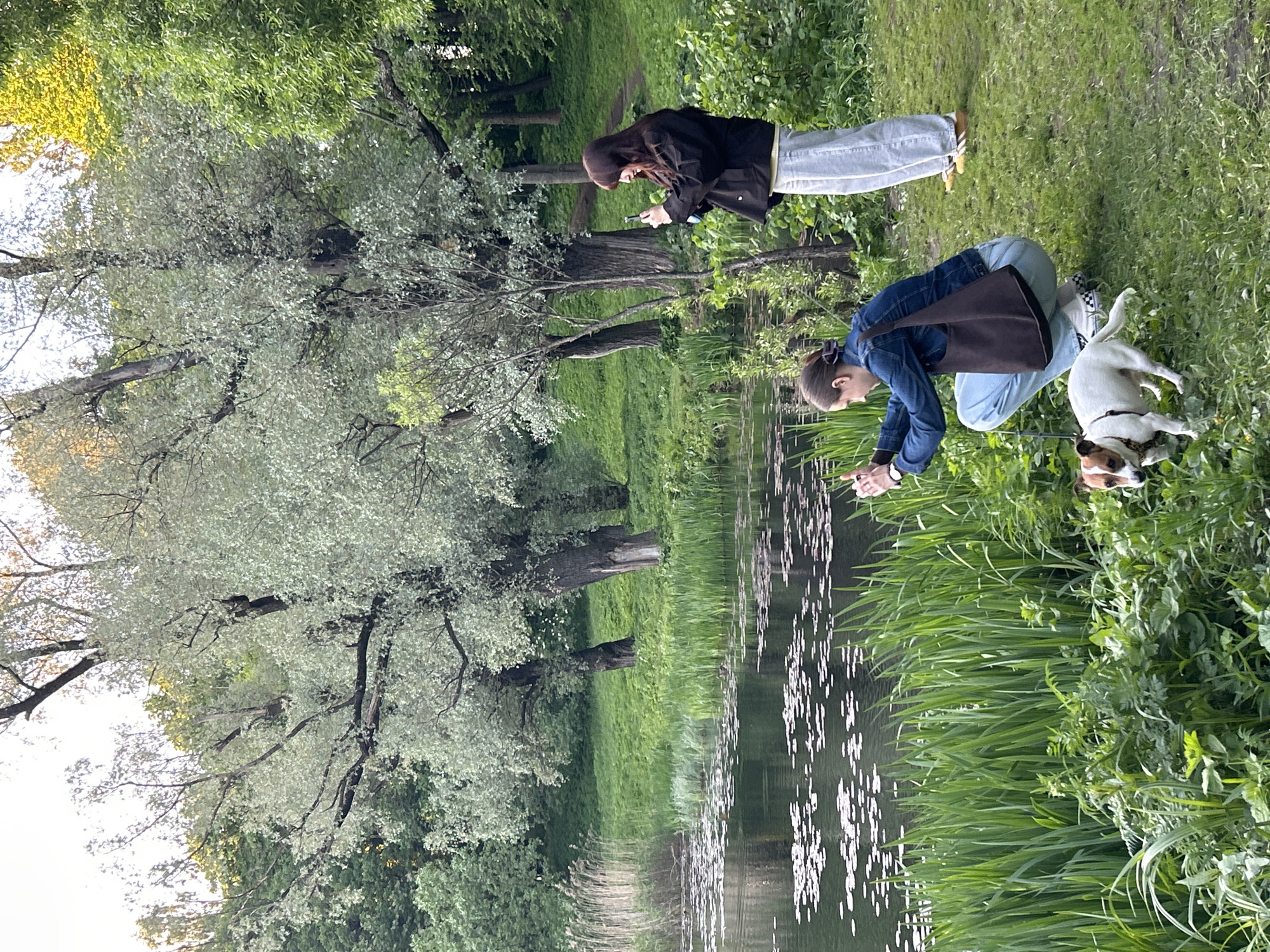 The scene depicts two women and a dog at the edge of a body of water, likely a canal or river, in Saint Petersburg, Russia. Lush greenery surrounds the water. One woman, wearing a black jacket and light-colored jeans, stands and appears to be looking at or interacting with something on her phone. The second woman is squatting down, wearing a denim jacket and light-colored jeans, with a brown bag over her shoulder. She seems to be interacting with the water, possibly touching it. A small dog stands near her, partially on the grassy bank and partially in the water. Trees with light green foliage are visible in the background. The water reflects the sky and the surrounding vegetation.