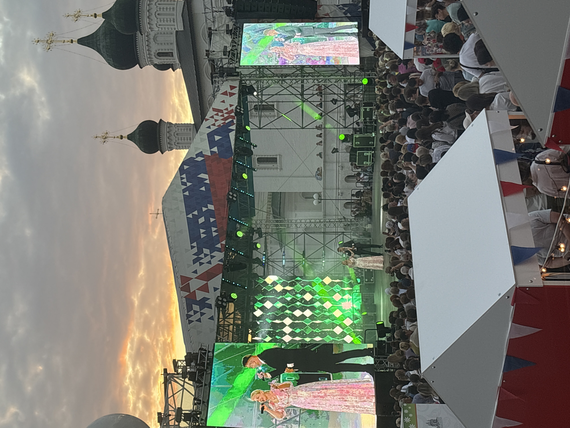 In Astrakhan, Russia, an outdoor concert is in progress at sunset. A large stage is set up with prominent video screens displaying performers. On the main screen, a male and female duo are singing, with the female in a shimmering pink gown and the male in a dark suit. A second, smaller screen to the right also shows a similar pair performing. A crowd of spectators fills the area in front of the stage, many holding up phones to capture the event. Behind the stage, the distinctive architecture of white buildings with green domes and golden crosses, characteristic of Orthodox cathedrals, is visible against the colorful twilight sky. Structures with white roofs and red and blue triangular banners are set up in the foreground, suggesting a festival or public gathering. The overall scene conveys a lively atmosphere of a cultural event.