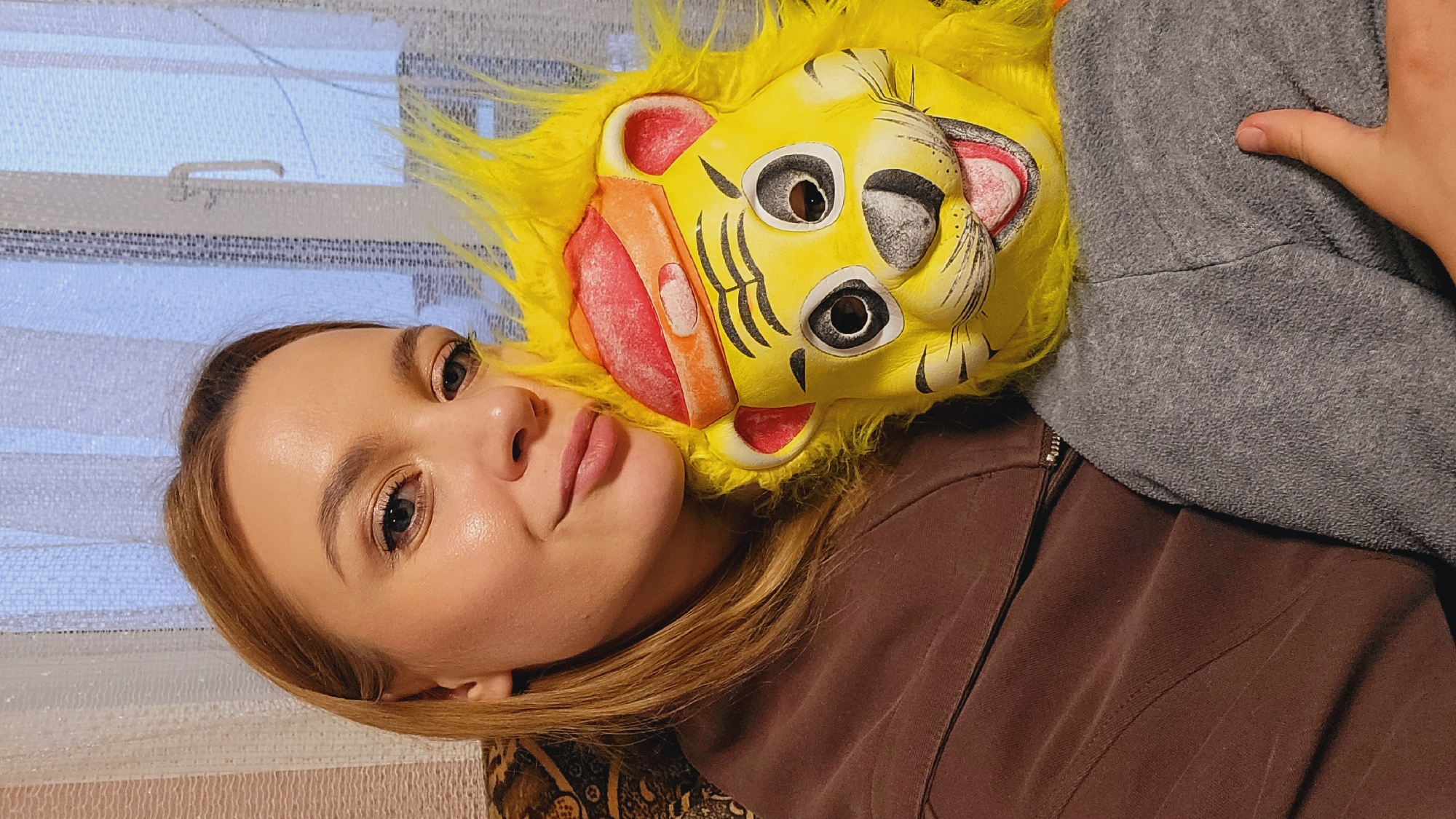 A woman with long, light brown hair is lying down indoors. Her head is tilted, and she's looking at the camera with a slight smile. A yellow lion mask with faux fur is lying next to her head, touching her face. She is wearing a brown shirt. A gray hooded sweatshirt partially covers the right side of the image, and a hand is resting on it. A window with a striped curtain is visible in the background. This scene is located in the city of Зипуново, Russia.