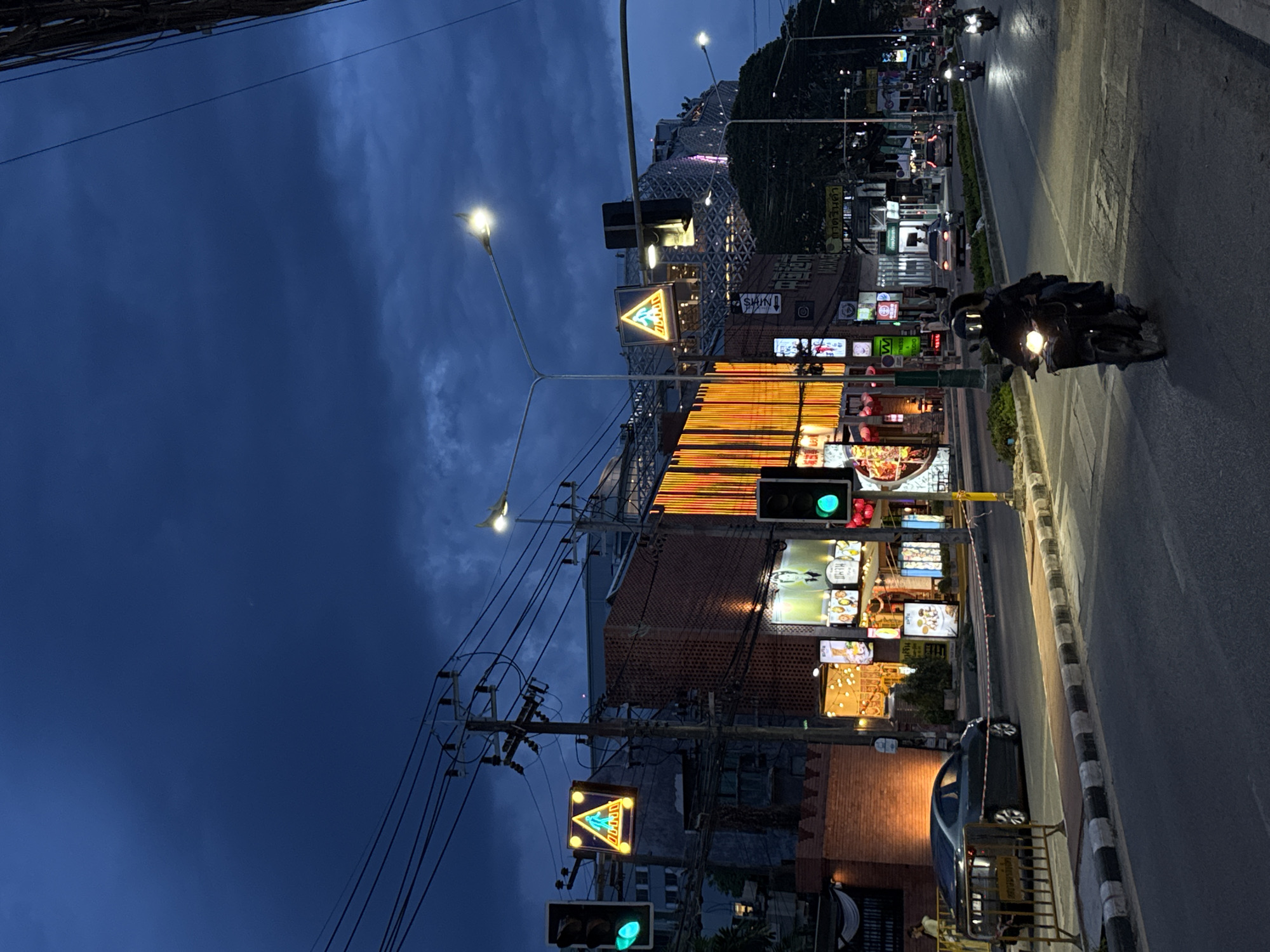 The image depicts a street in Chiang Mai City Municipality, Thailand, at night. The sky is a deep blue with some clouds. The street is illuminated by streetlights and neon signs from businesses lining the road. There are traffic lights showing green. Several motorbikes are on the road, moving along the right side of the frame. The buildings feature a mix of brick and modern designs. A large building with a geometrical, latticed façade is in the background, also illuminated. Power lines are visible, crisscrossing the sky. Pedestrian crossing signs are also present. A parked car is on the right side of the road, near the curb. The overall scene is a vibrant urban environment at night.