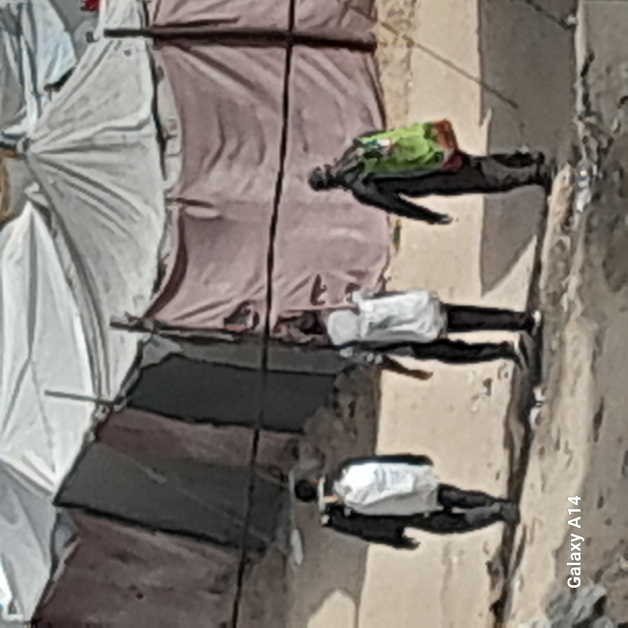 The image shows a street scene in an urban environment, potentially in an area of temporary housing or refugee camps. Three figures are visible, lying on the ground in the right side of the image. They appear to be dressed in clothing consistent with civilian attire. Adjacent to the figures are makeshift shelters constructed from cloth or tarpaulin. The ground is barren and dusty. The orientation of the scene is atypical, with the bottom of the image containing text "Galaxy A14".