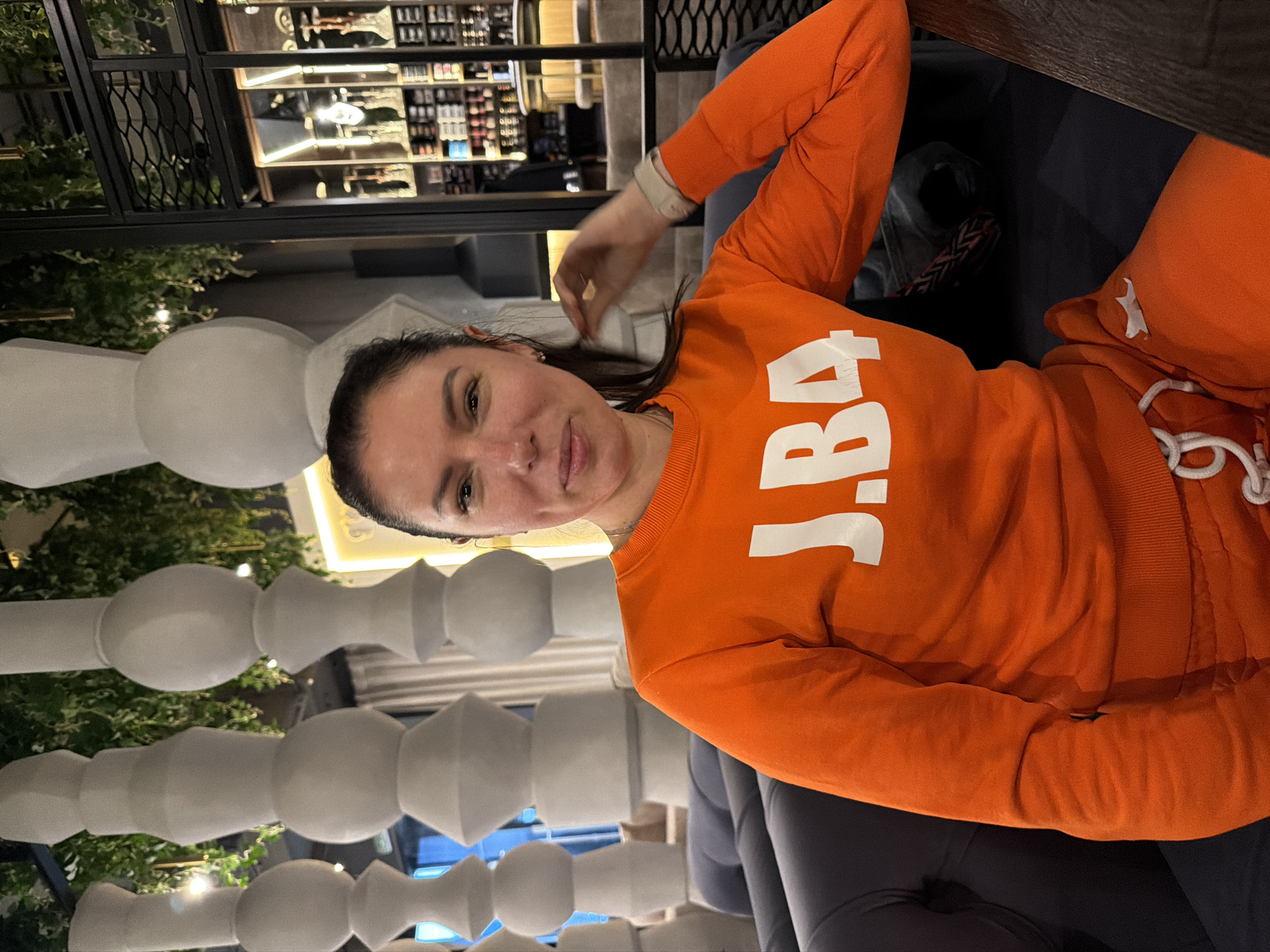 A woman is sitting in an armchair. The woman is wearing an orange sweatshirt with the letters "J.BA" in white. She is looking at the camera.
In the background, there is a bar with bottles on shelves. There are also white ornamental balusters in the background.
The image was taken in Krasnoyarsk, Russia.