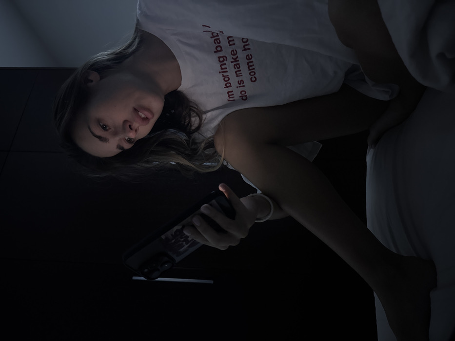 A young woman reclines in bed, bathed in the glow of a cell phone she holds in her hand. She wears a white t-shirt with the phrase "I'm boring baby/ do is make me/ come home" printed on it. Her gaze is directed at the viewer. The background is a dimly lit room with a dark-colored wardrobe. A partially visible white bed sheet covers the bed.