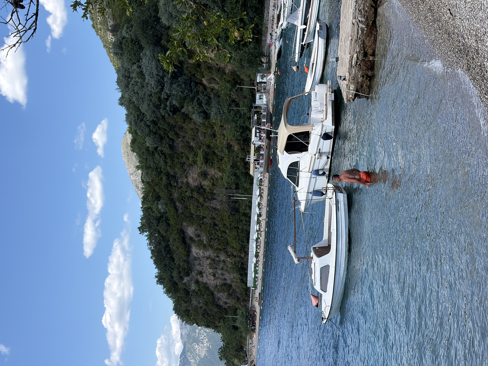 In Lepetane, Montenegro, a man in orange shorts stands in clear blue water near a small white boat. The boat, with a covered cabin, is moored next to other similar vessels. Beyond the boats, a pebble beach is lined with rows of white umbrellas and lounge chairs, suggesting a resort or public beach area. In the background, a lush green hillside rises steeply, dotted with trees and some rocky outcrops. The sky above is a bright blue, with scattered white clouds. The scene captures a sunny day by the Adriatic Sea, with a focus on leisure and the coastal landscape.