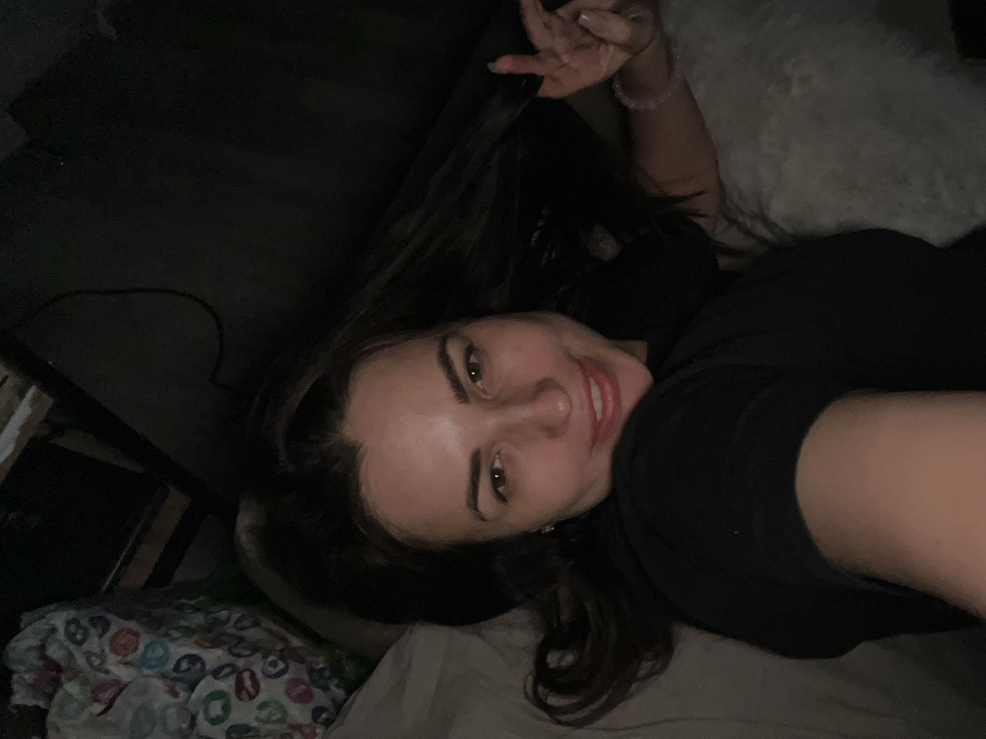 A woman lies in bed, smiling and looking at the camera. She is wearing a black shirt and has dark hair. She is making a peace sign with her fingers. The background is dark, with a wooden object to the left. The location appears to be indoors.