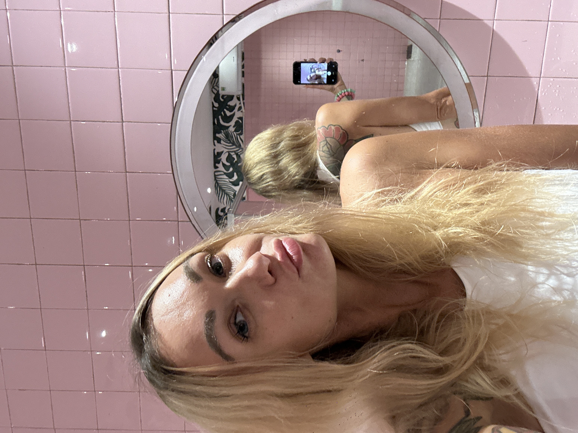 A blonde woman is featured in a mirror selfie, with her head slightly tilted and her gaze directed towards the camera. She has blue eyes, full lips, and a somewhat pouty expression. Her long blonde hair frames her face and flows over her shoulders. She is wearing a white top, and tattoos are visible on her arms, including a floral design on her upper left arm.
The setting appears to be an indoor space, likely a bathroom or vanity area, characterized by square pink tiles covering the walls. A prominent feature is a round mirror with an illuminated white frame, positioned behind the woman's right side. The reflection in this mirror shows the woman's back and arm, holding a smartphone to capture the photo. Colorful beaded bracelets adorn her wrist in the reflection. Intriguingly, the smartphone screen itself reflects the scene, creating a "mirror selfie within a mirror selfie" effect. Behind the woman's reflection in the mirror, a black and white tropical leaf pattern is visible, suggesting a decorative element like wallpaper or a shower curtain. The lighting is artificial, emanating from the mirror's illumination. No visible text is present. The context provided is Tien Thanh Ward, Vietnam.