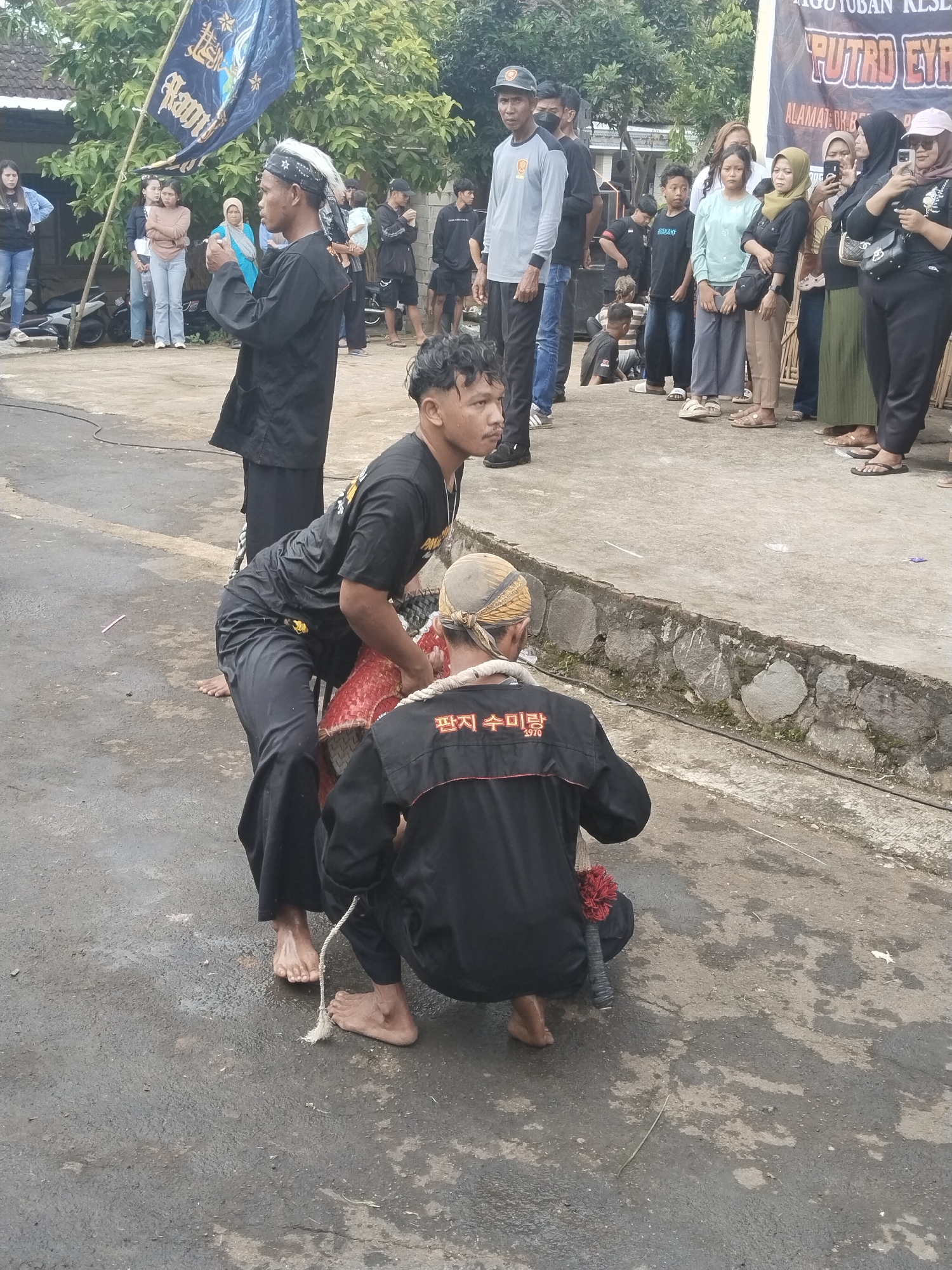 In Pendem, Indonesia, a group of people are gathered on a paved surface, likely a street or plaza. Two individuals, dressed in black attire with headbands, are interacting with a red drum-like object. One of the individuals is kneeling, while the other is leaning over the object. Both appear to be barefoot. A third individual stands to the left, also in black attire and a headband, with his arms folded, observing the scene. In the background, a larger group of people is watching the event. A flag with writing on it is positioned to the left. A banner with text is visible in the background on the right. The environment appears to be an outdoor setting with trees and buildings visible in the distance.