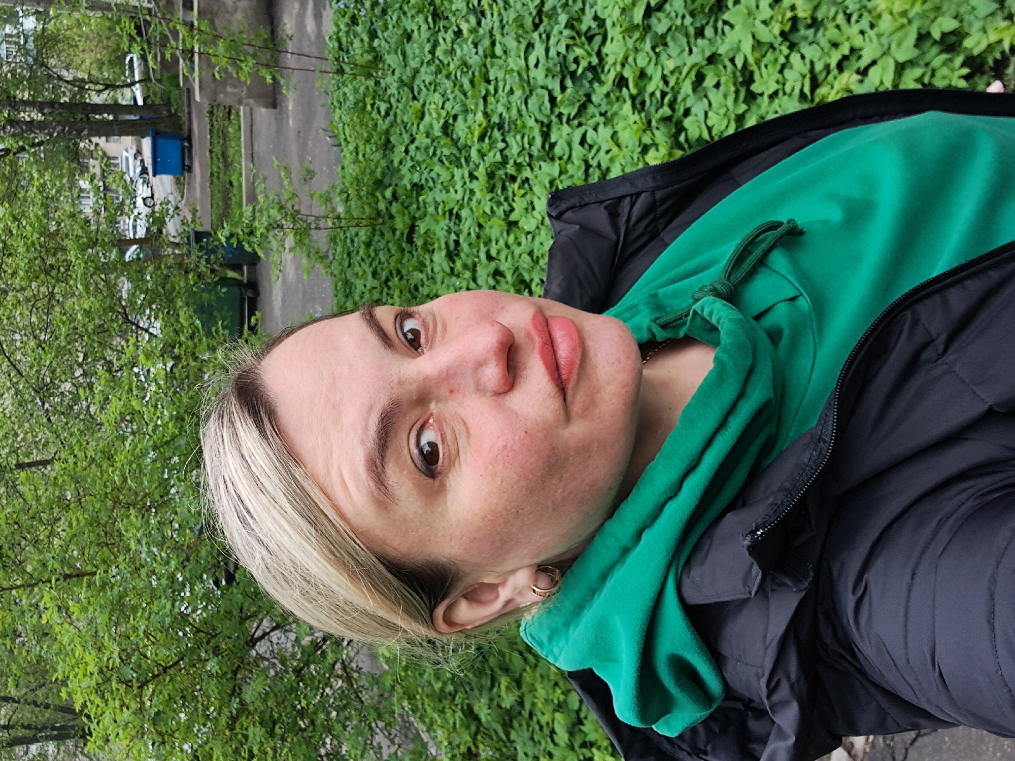 A woman is taking a selfie outdoors. She has blonde hair, fair skin, and is wearing a green hooded sweatshirt and a black jacket. The background consists of green vegetation, including trees and ground cover, and a glimpse of a street with cars and buildings. The location is Elektrostal, Russia.