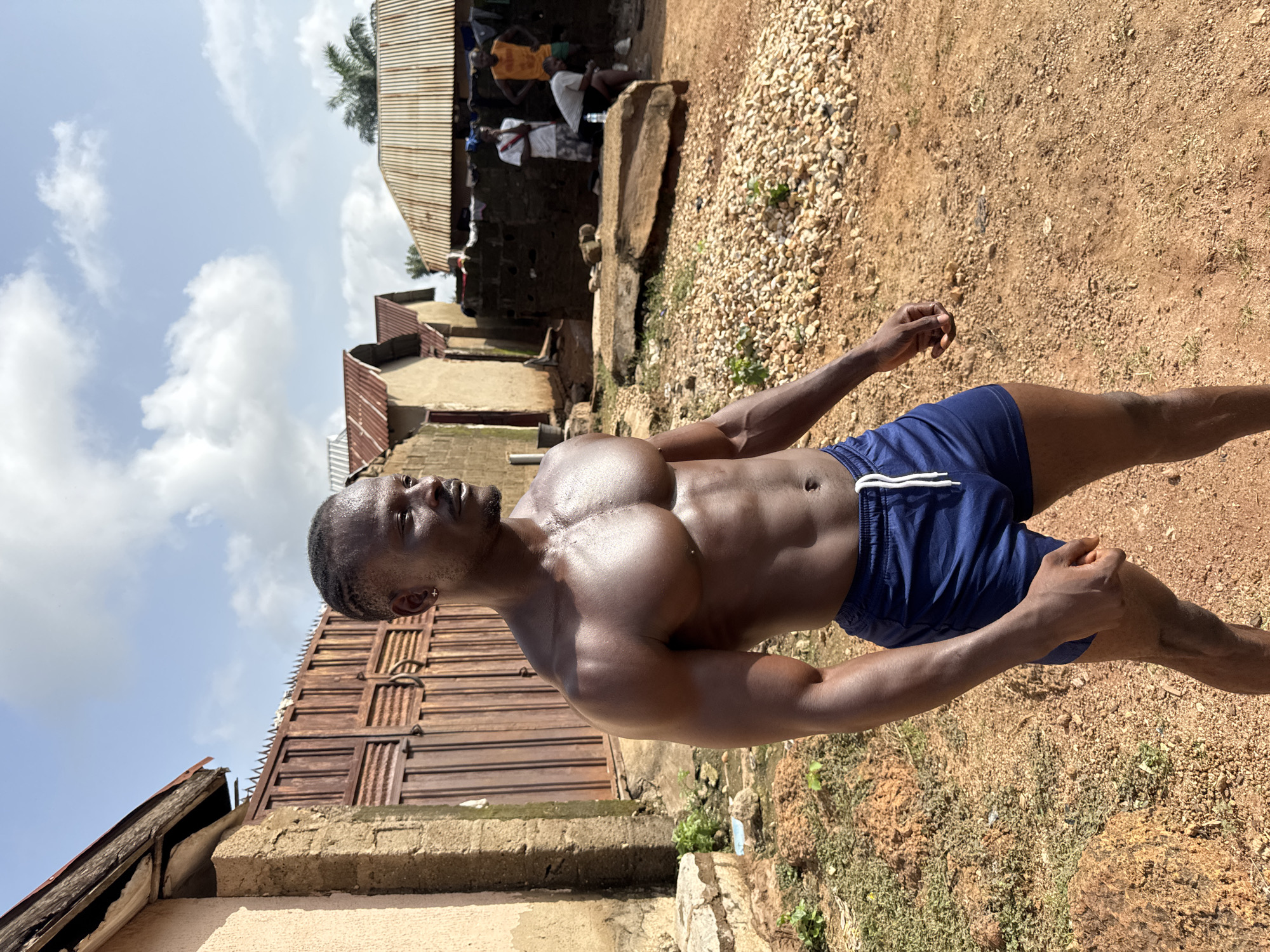 A man stands shirtless in Akwanga, Nigeria, revealing a muscular physique. He wears blue shorts with white drawstrings. His skin glistens, possibly due to sweat or oil. He is surrounded by a typical Nigerian landscape: houses with corrugated iron roofs and weathered walls, a gated entrance, and a gravel and dirt ground cover. Other people are visible in the background near a structure with a corrugated roof. The sky is partly cloudy.