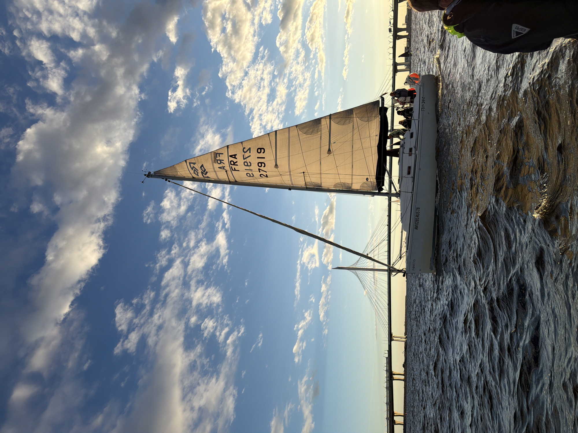 The image captures a sailboat navigating a body of water, likely a gulf or bay, with a large cable-stayed bridge in the background. The sky is blue with scattered white clouds, and the sunlight suggests it is either early morning or late afternoon. On the sail of the sailboat, the letters "FRA" are visible, along with the number "27919". The sailboat appears to have several people on board. The bridge in the background is a prominent structure, indicative of an urban environment. Given the presence of the distinctive bridge and the water body, this scene is located in Saint Petersburg, Russia, likely on the Gulf of Finland, with the bridge being the Western High-Speed Diameter. The water is choppy, indicating some wind.