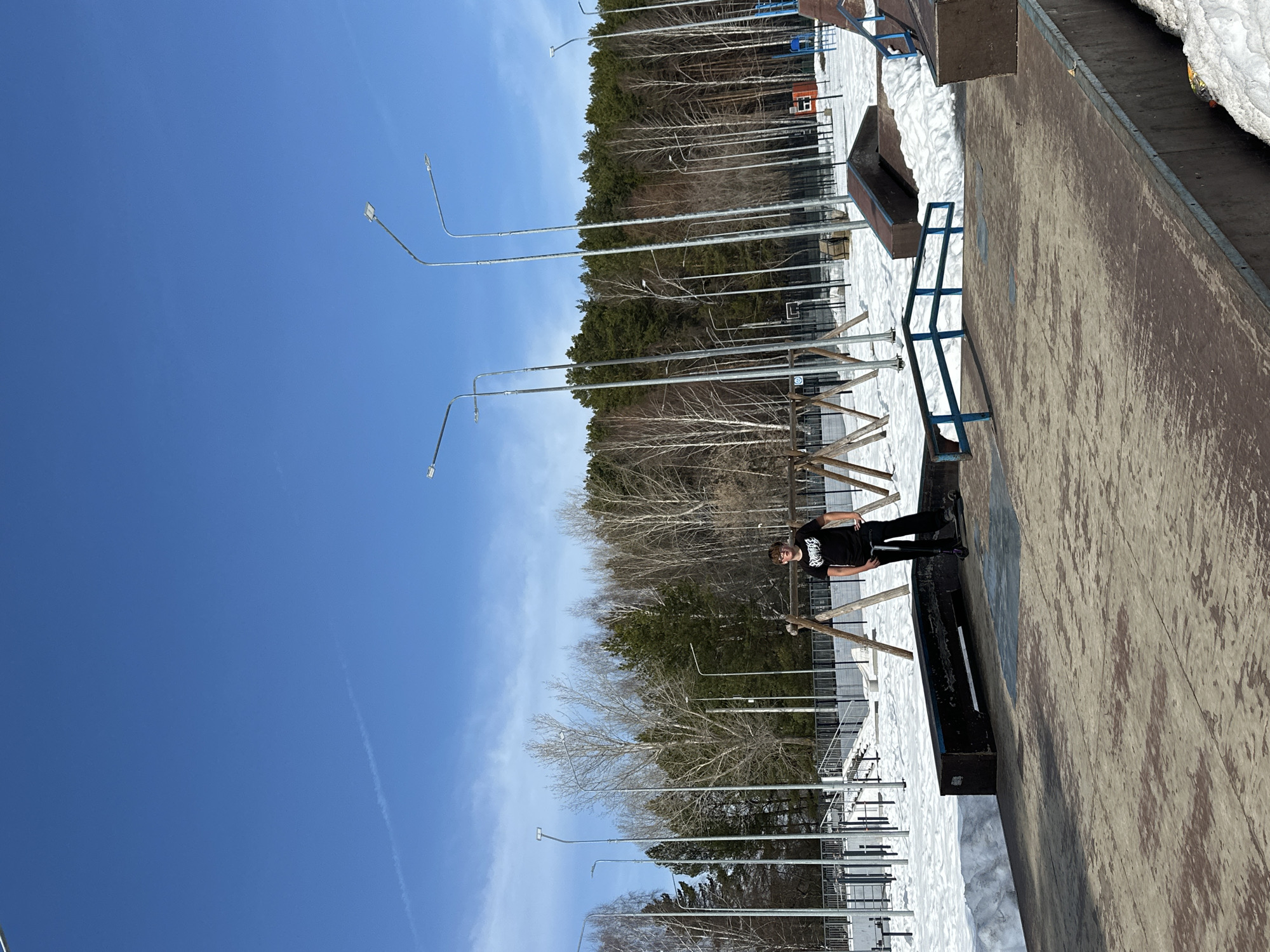The image depicts an outdoor scene, likely at a skate park in Chaikovsky, Russia. The sky is a clear blue with a contrail of white clouds. In the foreground and mid-ground, there is a concrete skate park structure, with a ramp and various obstacles. Patches of snow are visible around the skate park, suggesting either late winter or early spring. A young man, wearing a black t-shirt, black pants, and glasses is standing on a ledge, holding a scooter. Tall metal light poles line the area near the park. Behind the skate park, a dense forest of evergreen trees extends to the horizon. The overall environment suggests an urban setting near a natural forested area.